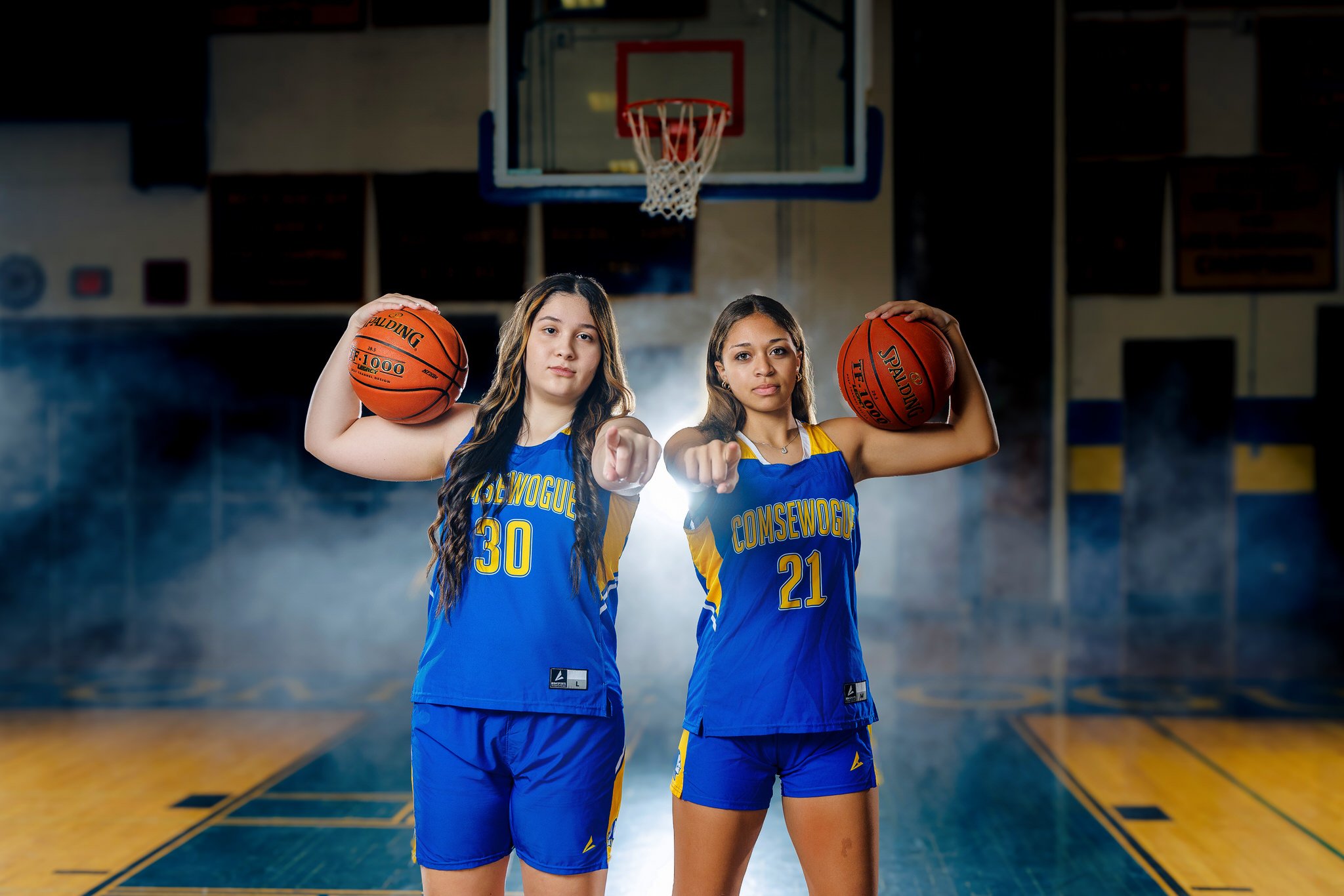 Two female basketball players in blue and yellow uniforms, posing with basketballs on a basketball court, pointing towards the camera.