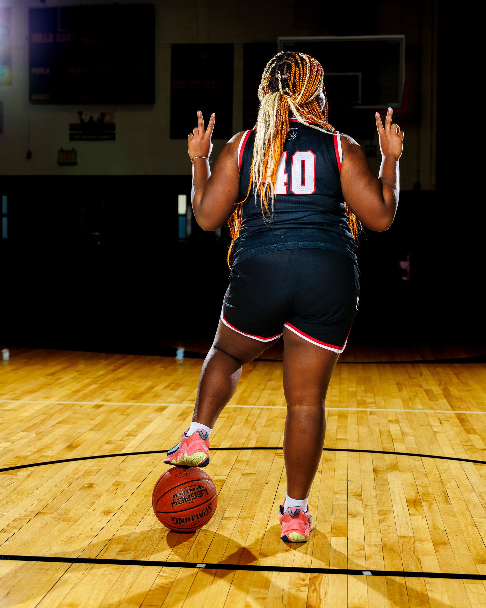 A female basketball player is standing on a basketball court with her back to the camera. She has long braids and is wearing a black jersey with the number 10, black shorts with red and white trim, and pink athletic shoes. She is balancing a basketba