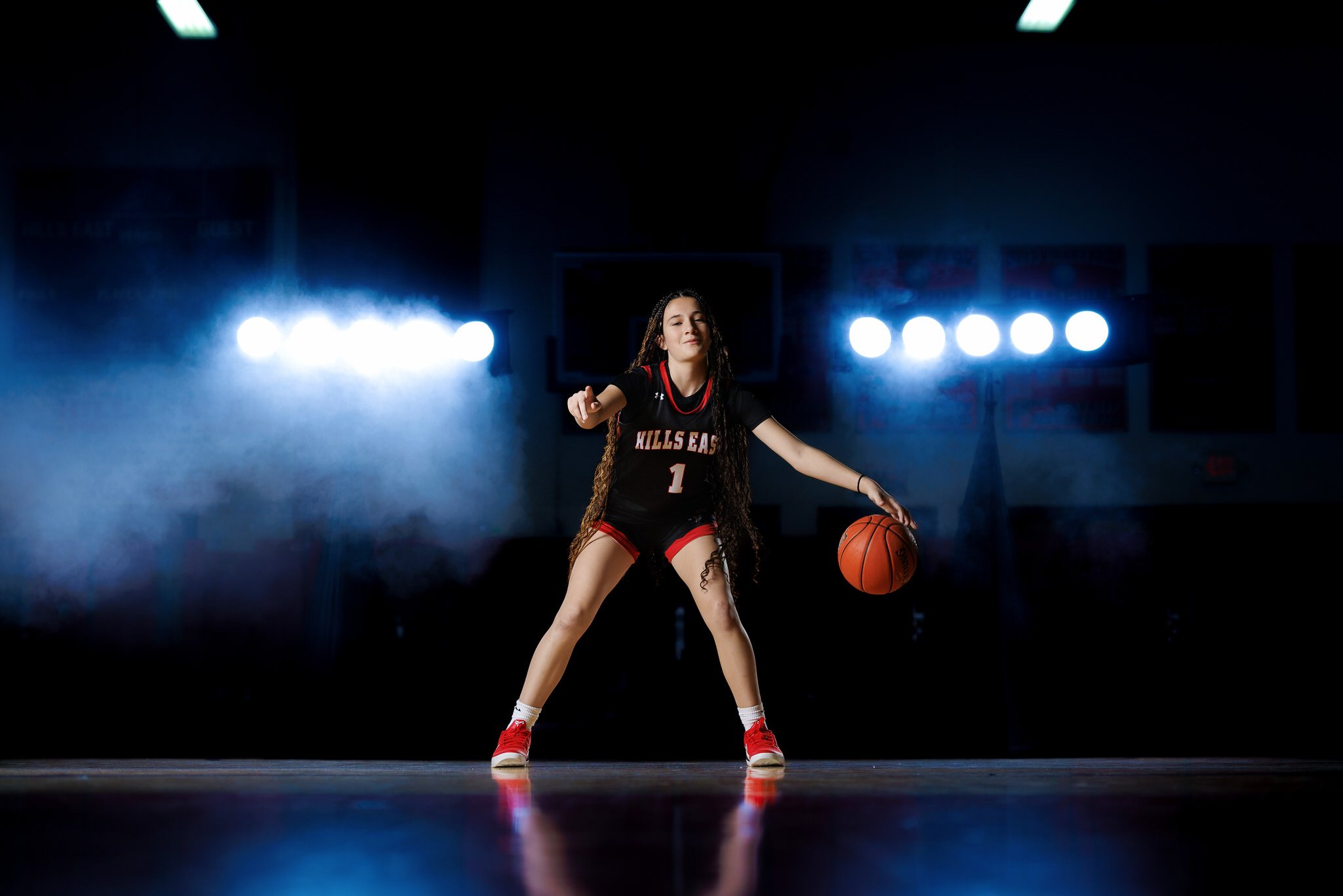 A young female basketball player practicing dribbling in a dark gym with spotlights, wearing a black and red jersey that reads 'Hills East' and the number 1.