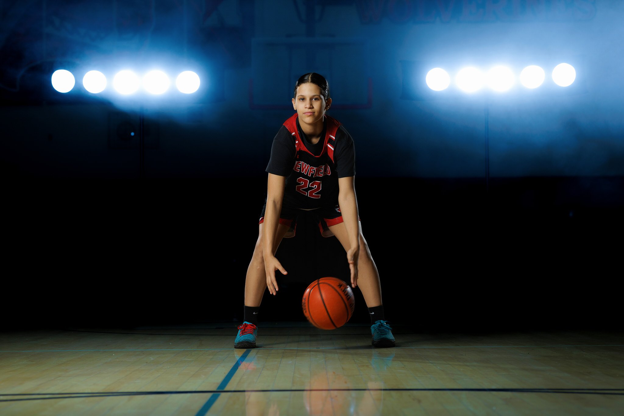 A young female basketball player in a black and red uniform, crouching with a basketball on the court, in a gym with bright overhead lights.