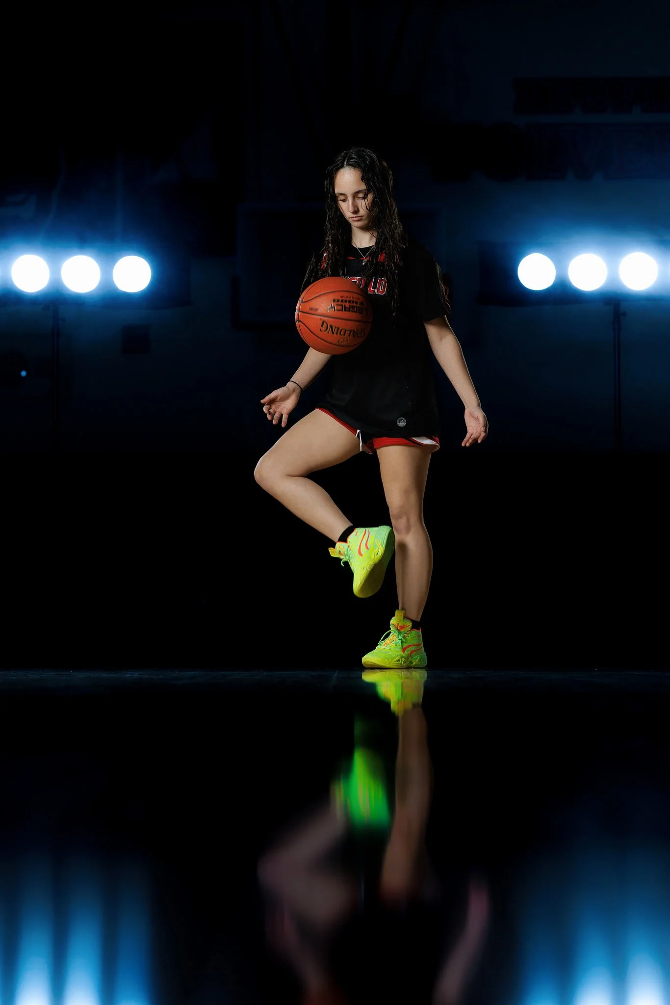 Young woman in black and red sportswear balancing a basketball on her thigh in a dark gym with bright lights in the background.