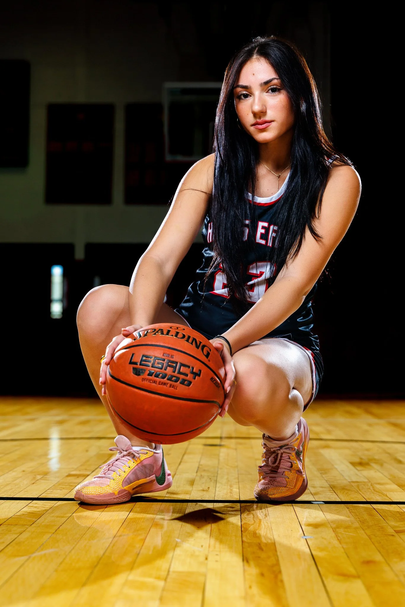 A young woman in a basketball uniform squatting on a basketball court, holding a basketball with both hands, with a focused expression.