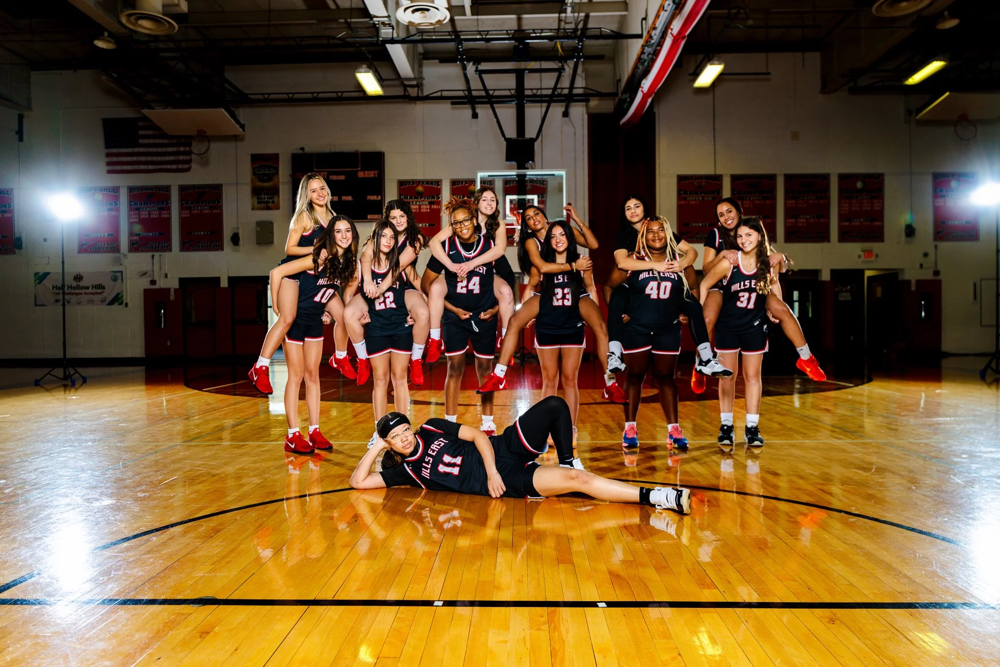 A girls volleyball team in uniform posing and smiling on a basketball court in a gymnasium.