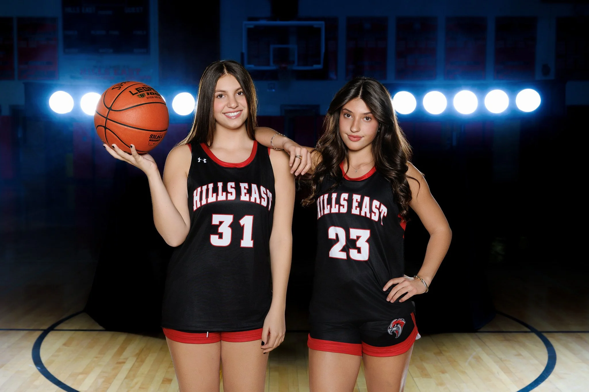 Two young women wearing black and red basketball jerseys with 'HILLS EAST' written on them, standing on a basketball court at night with bright lights in the background. One is holding a basketball.