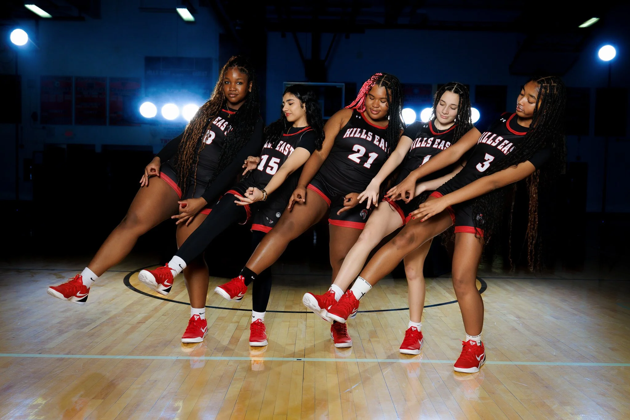 Five young women in black and red basketball uniforms standing in a gymnasium with a wooden floor and blue lighting in the background.