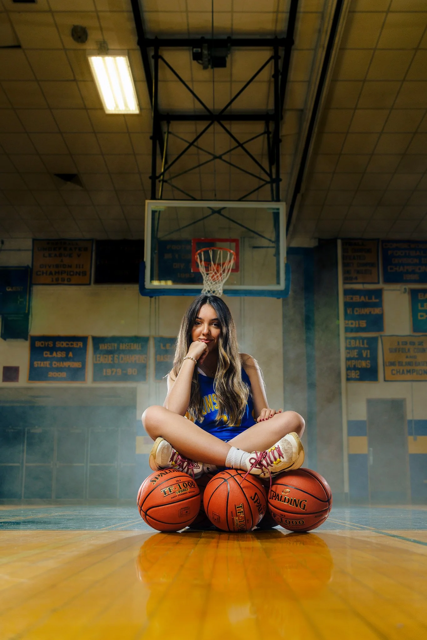 A young woman sitting cross-legged on a basketball court with three basketballs in front of her. She is wearing a blue sports jersey, sneakers, and has long, wavy hair. In the background, there is a basketball hoop and banners on the wall celebrating