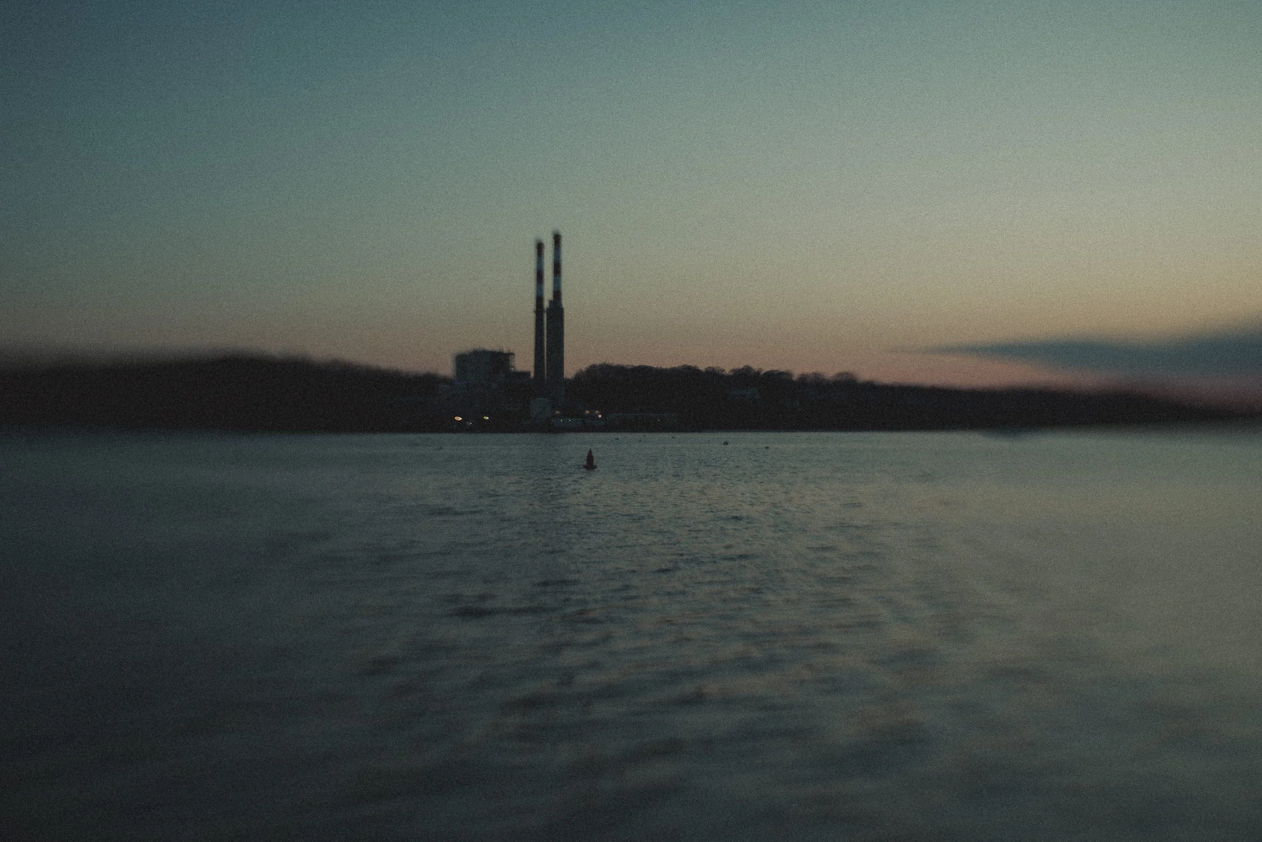 A body of water with a smokestack or industrial chimney in the distance on the horizon during dusk or dawn.