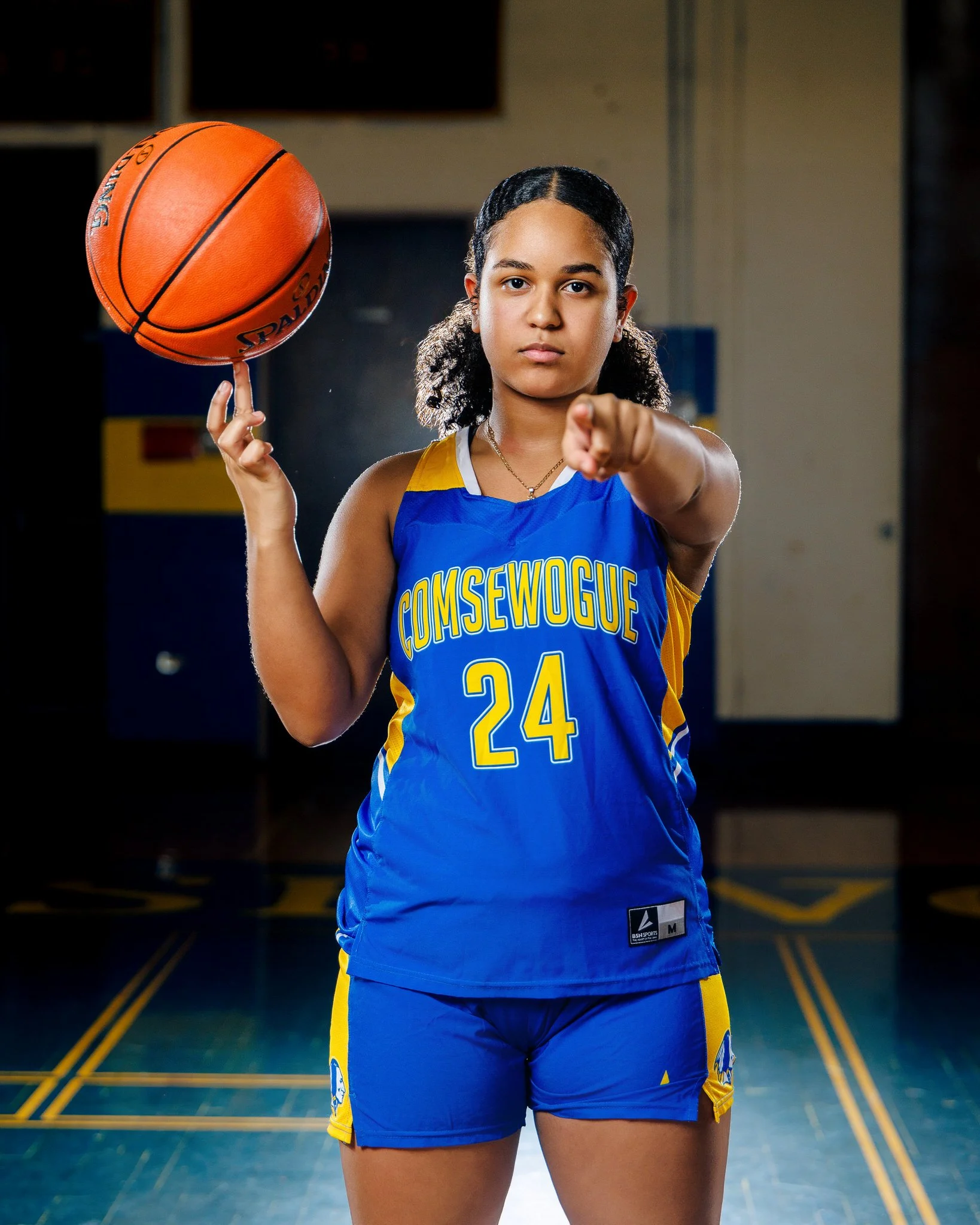 A young female basketball player in a blue and yellow jersey holding a basketball and pointing at the camera inside a gymnasium.
