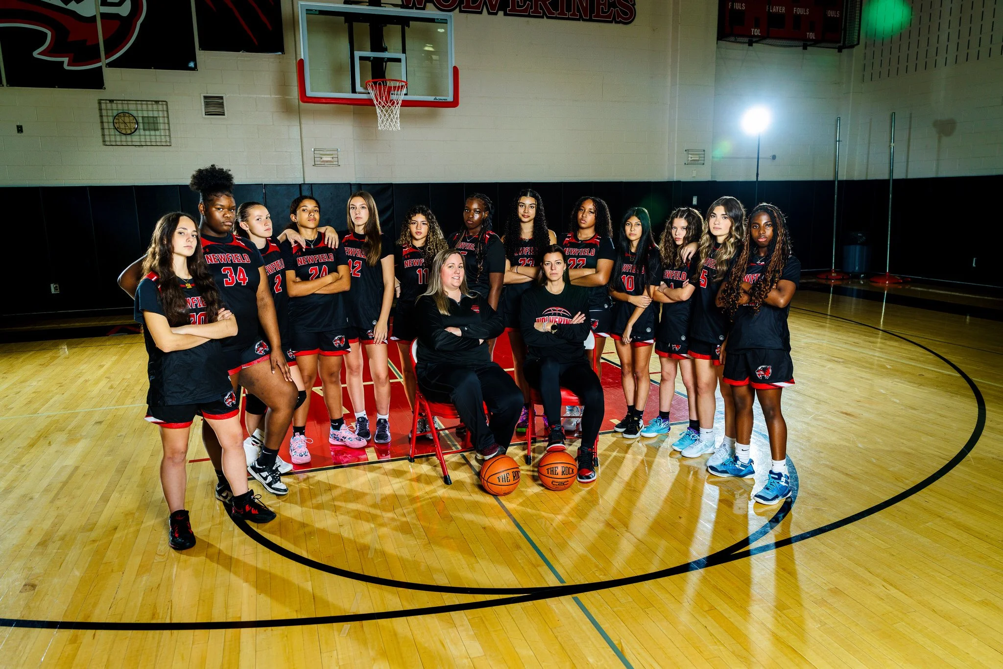 A girls' basketball team in matching black and red uniforms posing inside a gymnasium, with coaches seated in front and players standing behind, on a polished wood basketball court.