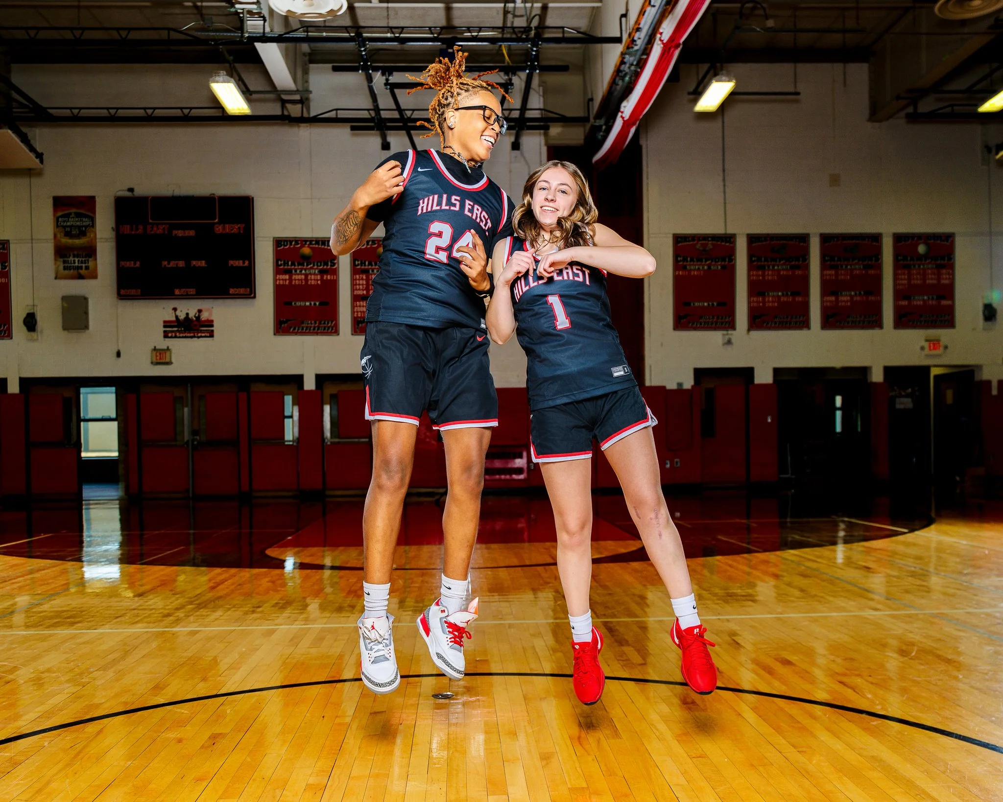 Two young female basketball players in navy blue jerseys and shorts jumping and smiling on a basketball court. One has dreadlocks and glasses, the other has curly hair. The court has a wooden floor and a gymnasium background with banners and a scoreb