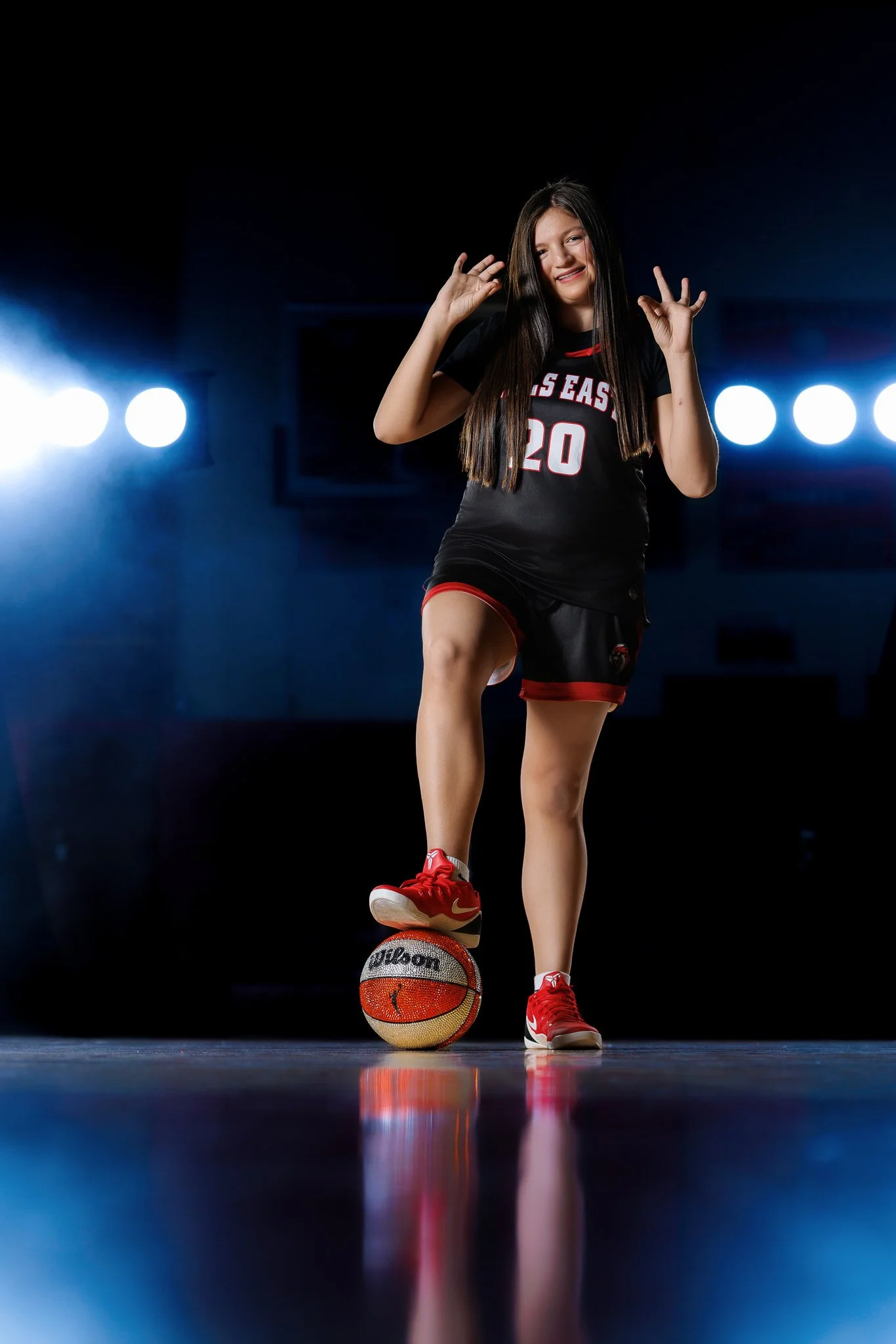 Young woman in basketball uniform smiling, balancing a basketball on her foot, with bright stage lights in background.