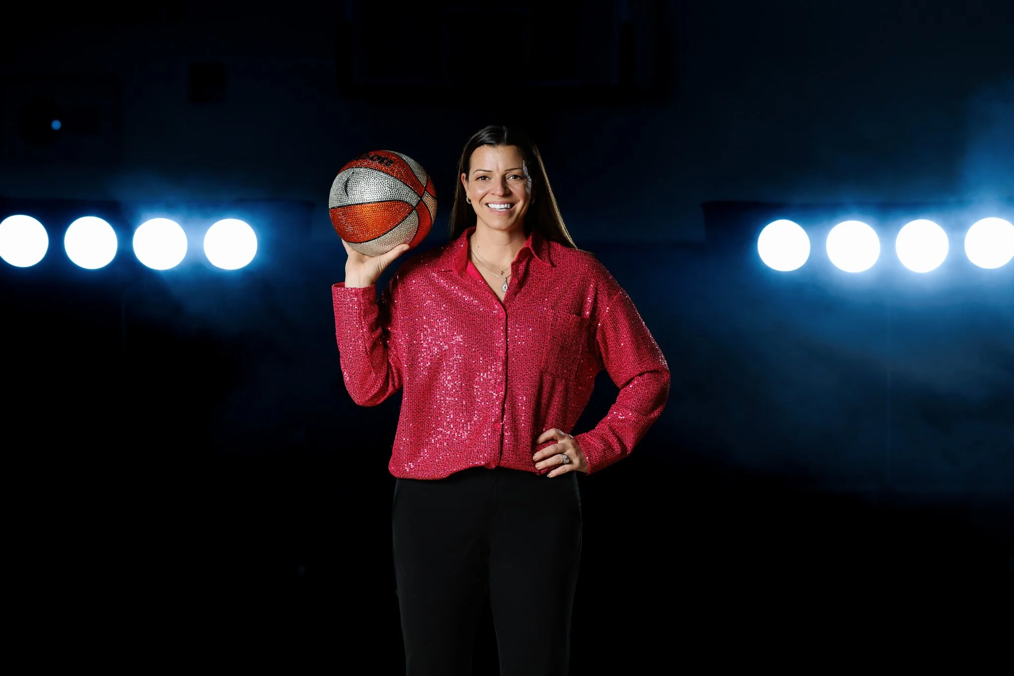 A woman smiling and holding a basketball with her right hand, wearing a pink sequin shirt, standing in front of bright stage lights.