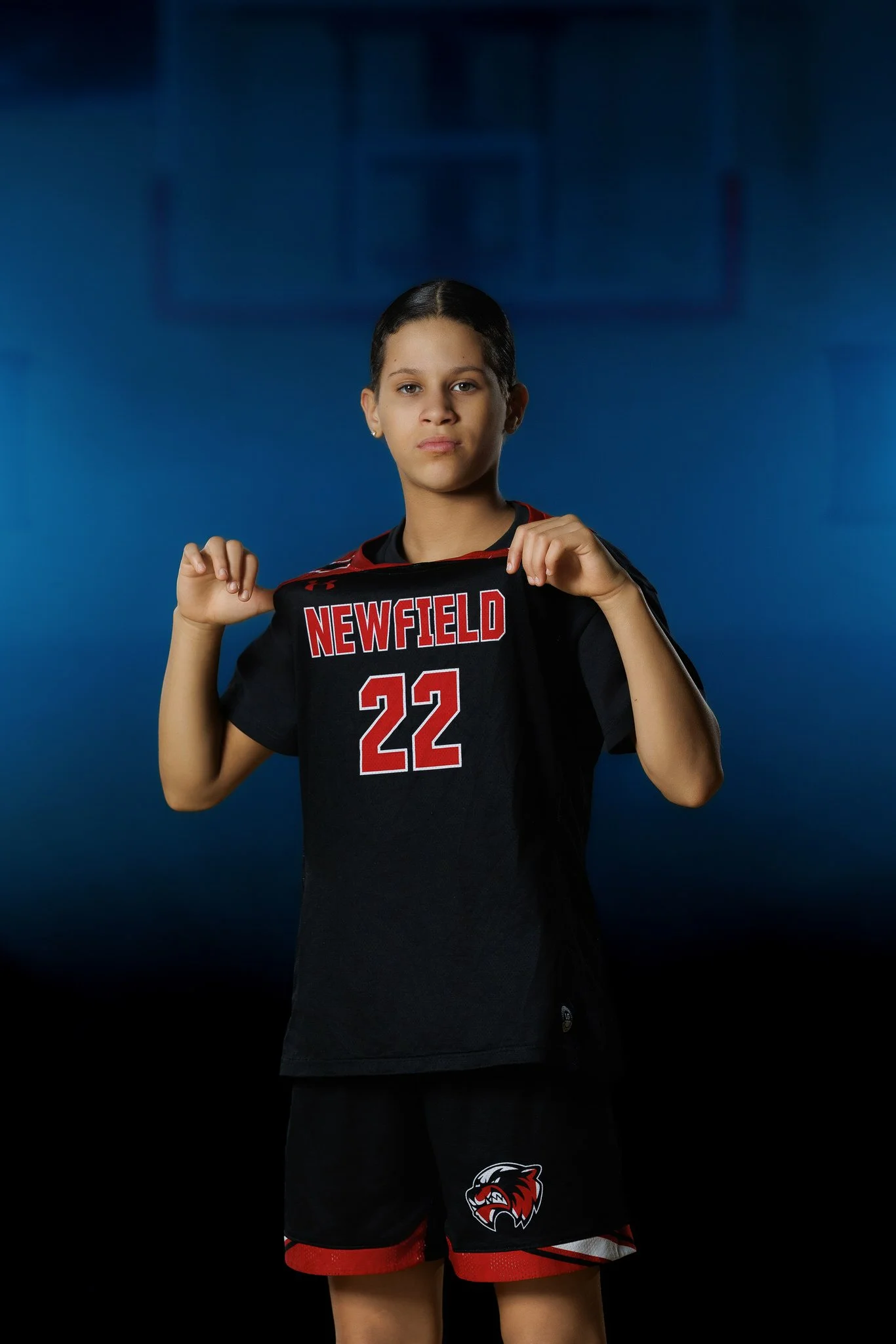 A young female athlete wearing a black and red sports uniform with the words "Newfield" and the number 22, holding up her jersey in front of a blue background.