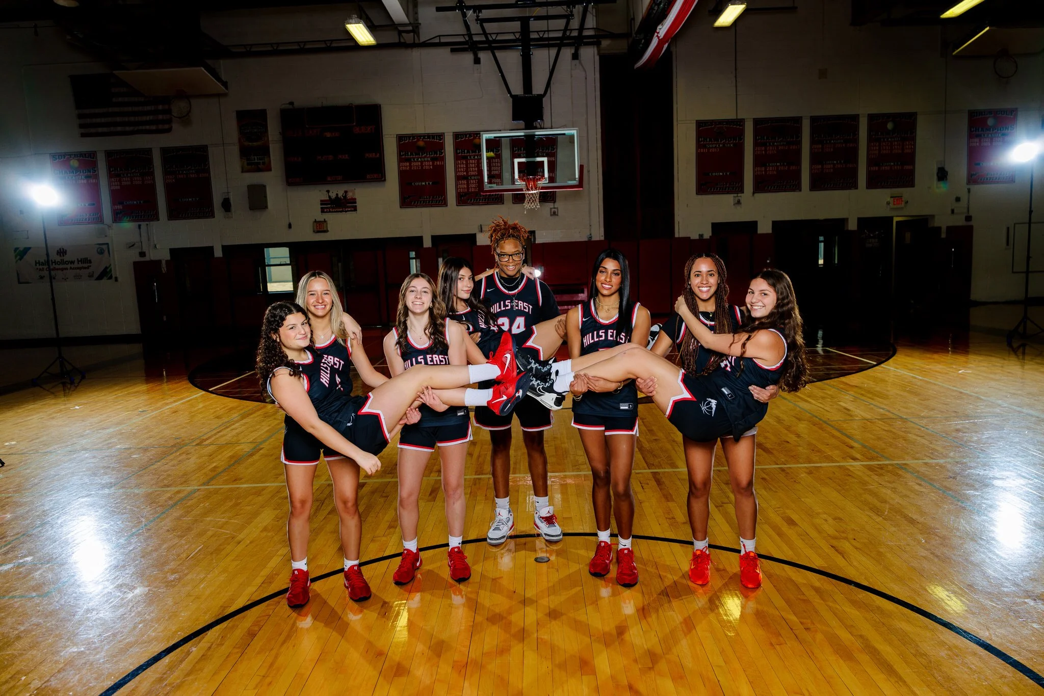 A group of young female basketball players in matching uniforms on an indoor basketball court, with one girl being held horizontally by the others.