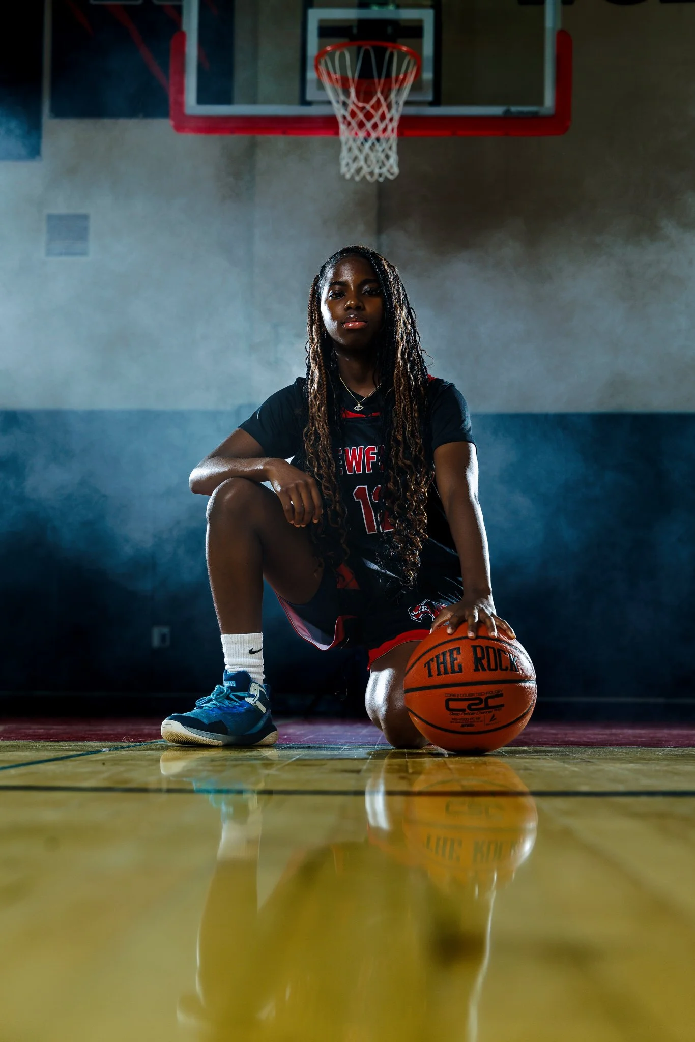 A female basketball player in a black uniform kneeling on a basketball court with one knee, holding an orange basketball with black text, in front of a hoop inside a gym.