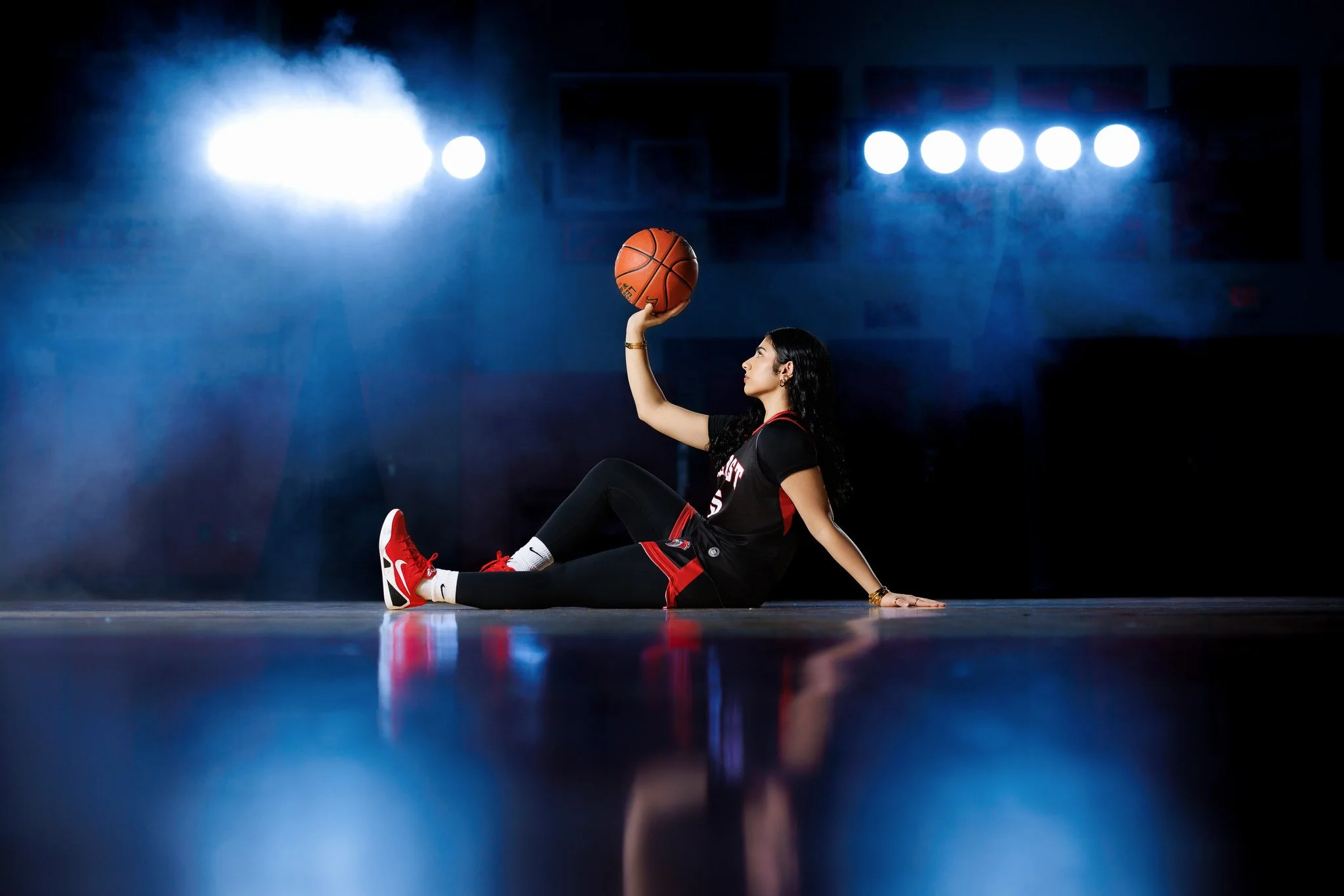 A female basketball player sitting on the court, holding a basketball above her head, with bright stadium lights in the background.