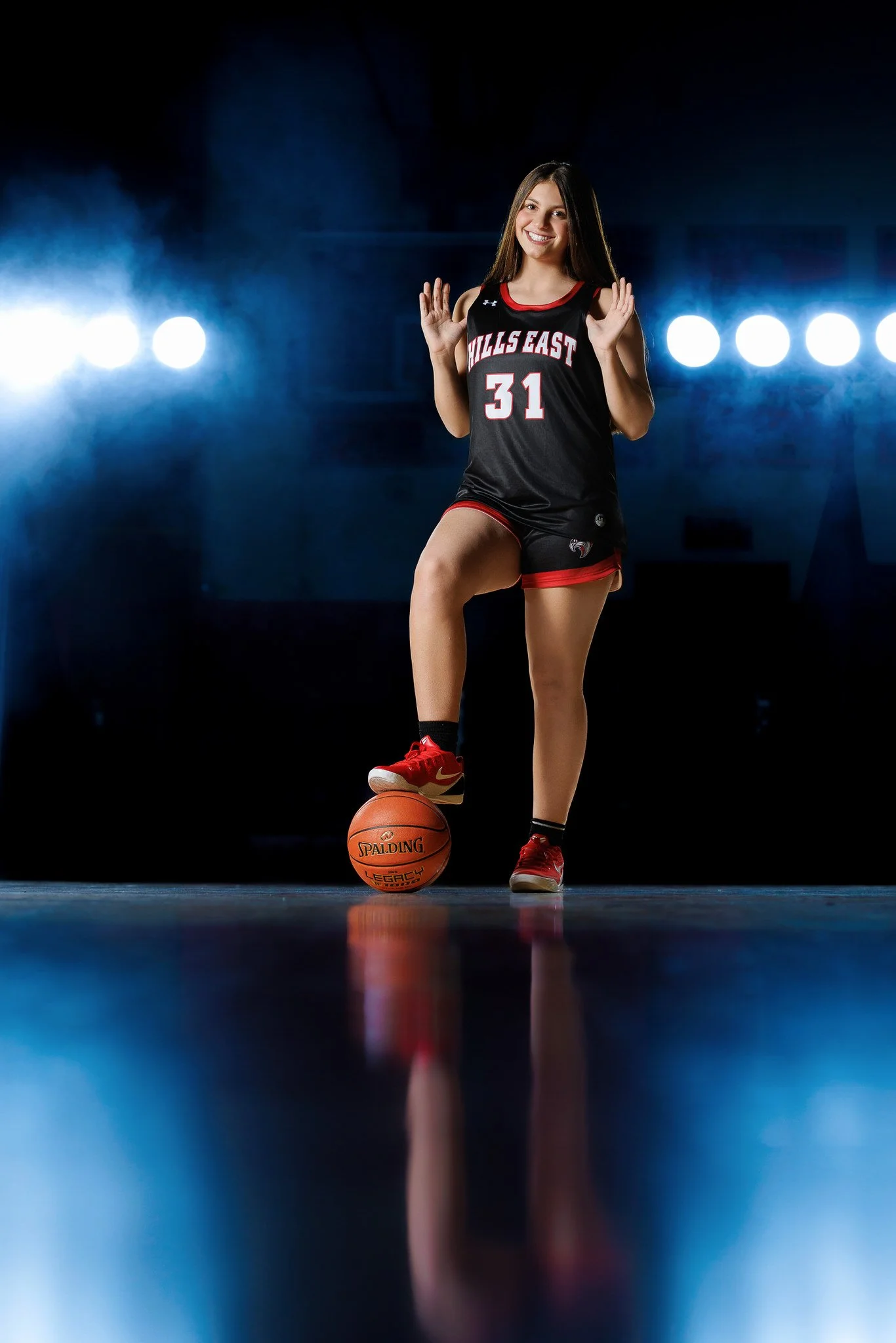 Young female basketball player in a black and red uniform with the number 31, standing on a basketball court with one foot on a basketball, smiling and waving, with bright lights in the background.
