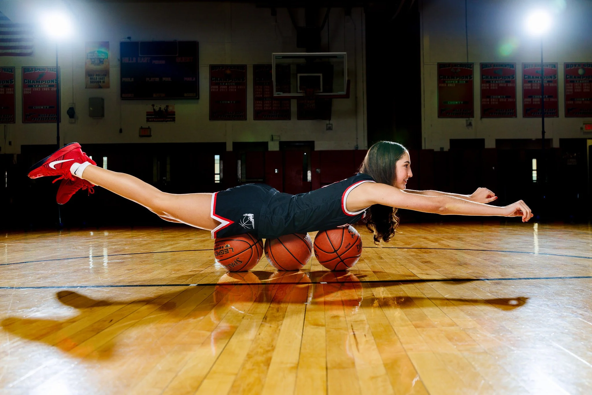 A girl in a basketball uniform balancing on three basketballs while stretching her arms forward in a gymnasium.