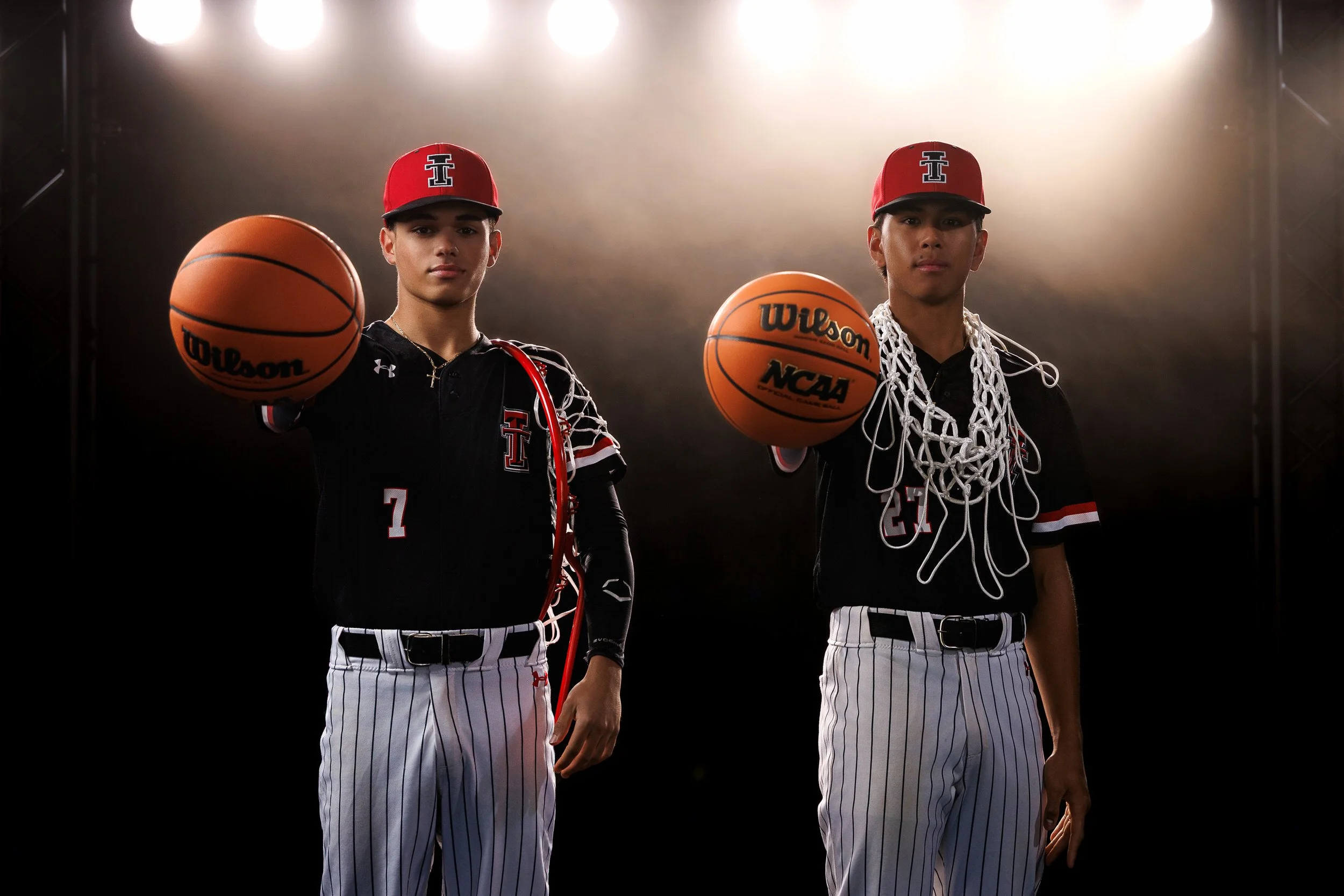 Two young male baseball players in black jerseys with white pinstripe pants and red baseball caps, holding basketballs, standing in a studio with bright lights behind them, one basketball has a net draped around his neck.