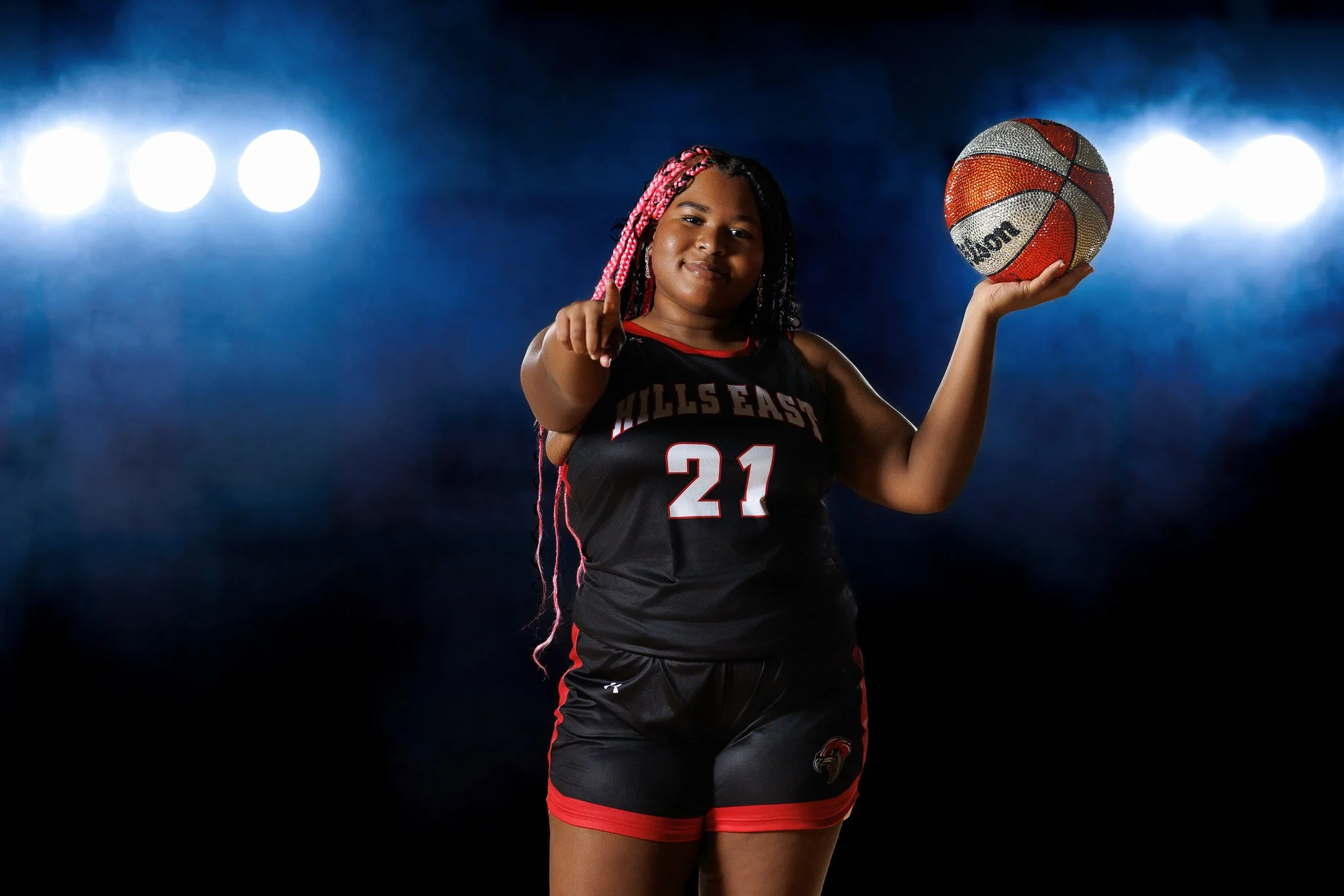 A young girl in a black and red basketball uniform in a sports studio, holding a basketball and pointing forward.