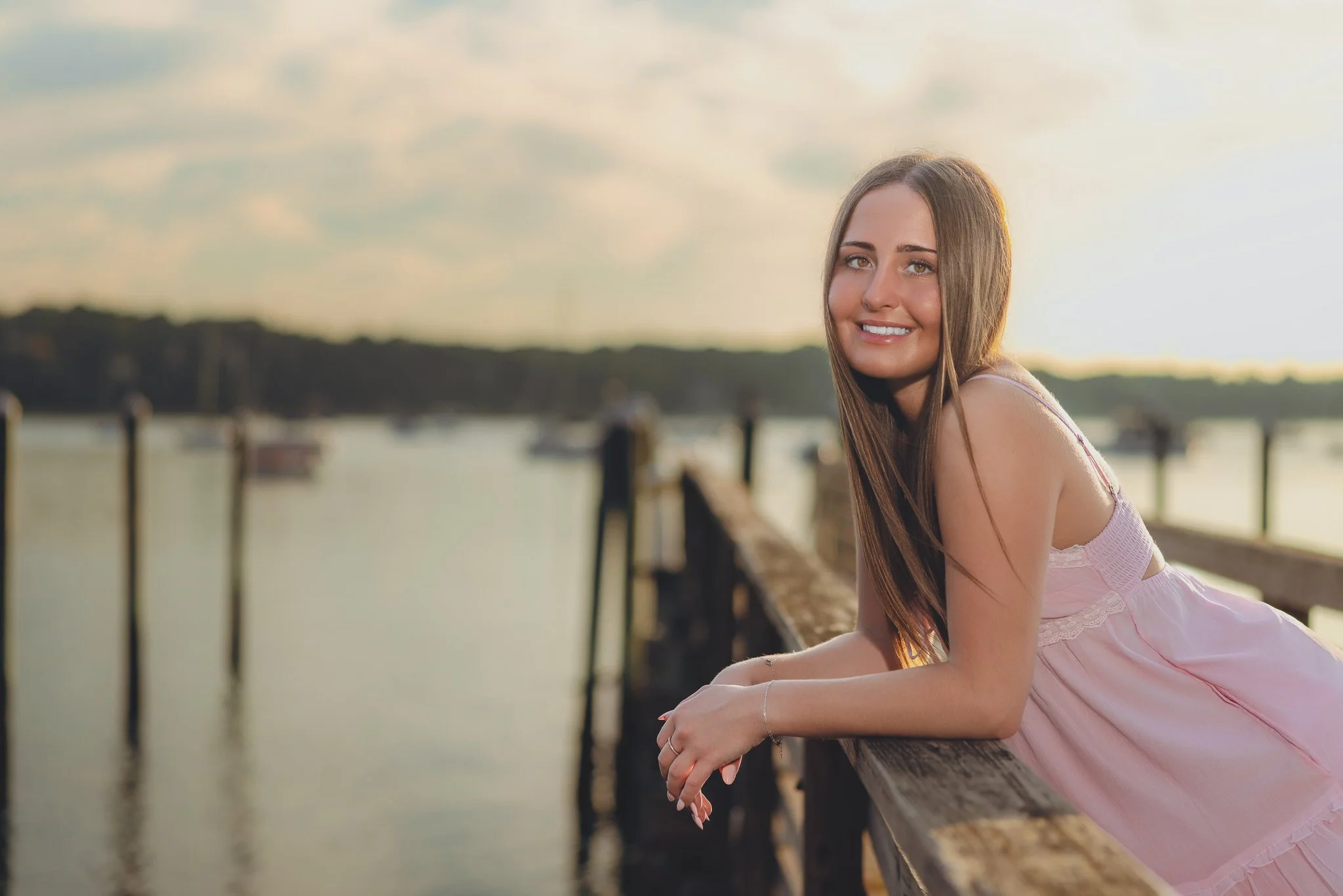 A young woman with long brown hair wearing a pink dress, leaning on a wooden railing by a body of water during sunset, smiling at the camera.