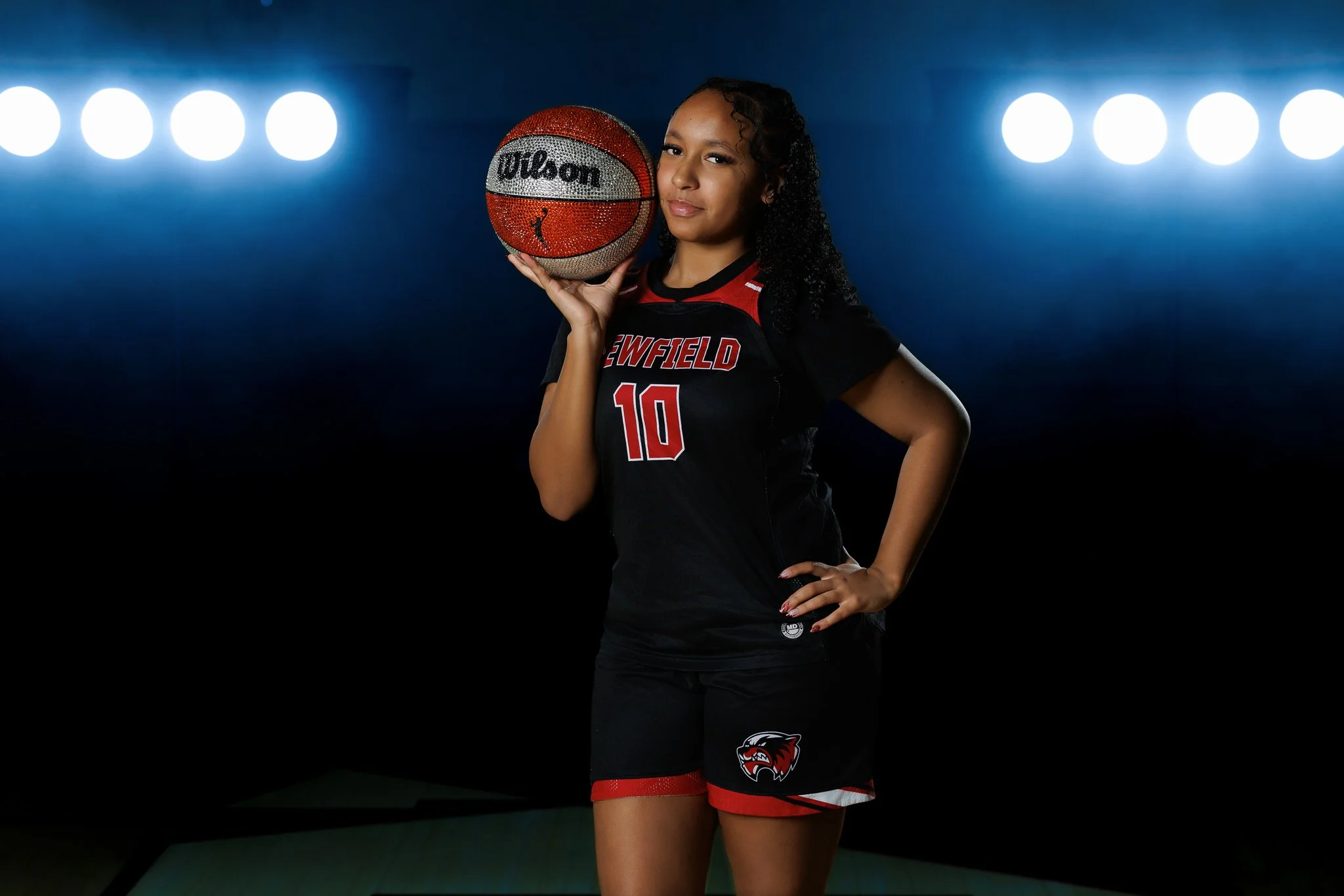 A young female basketball player with curly hair, wearing a black uniform with red and white accents, the number 10, and a team logo, holding a decorated basketball against her right shoulder, standing in a dark studio with bright blue lights in the 