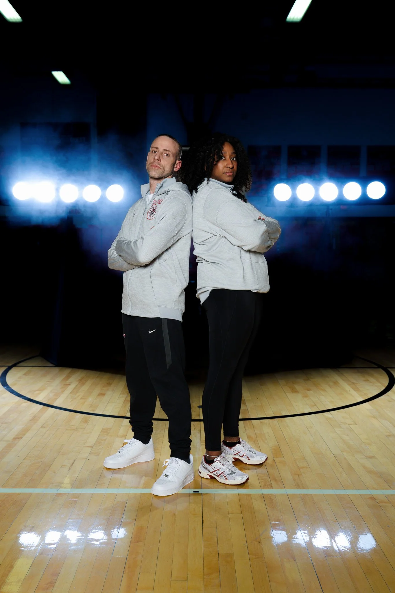 Two people, a man and a woman, standing back-to-back on a basketball court with their arms crossed, facing forward. They are wearing light gray hoodies and athletic pants, with sneakers. Bright lights and a smoky background are visible behind them.