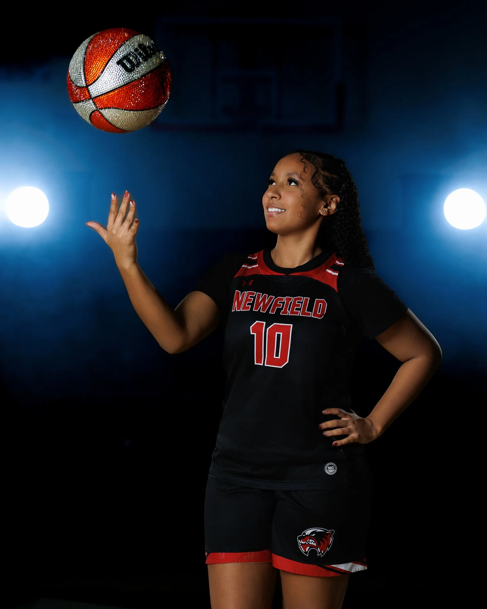 A female volleyball player wearing a black and red uniform with the number 10 on it, standing in a gymnasium, tossing a volleyball into the air.