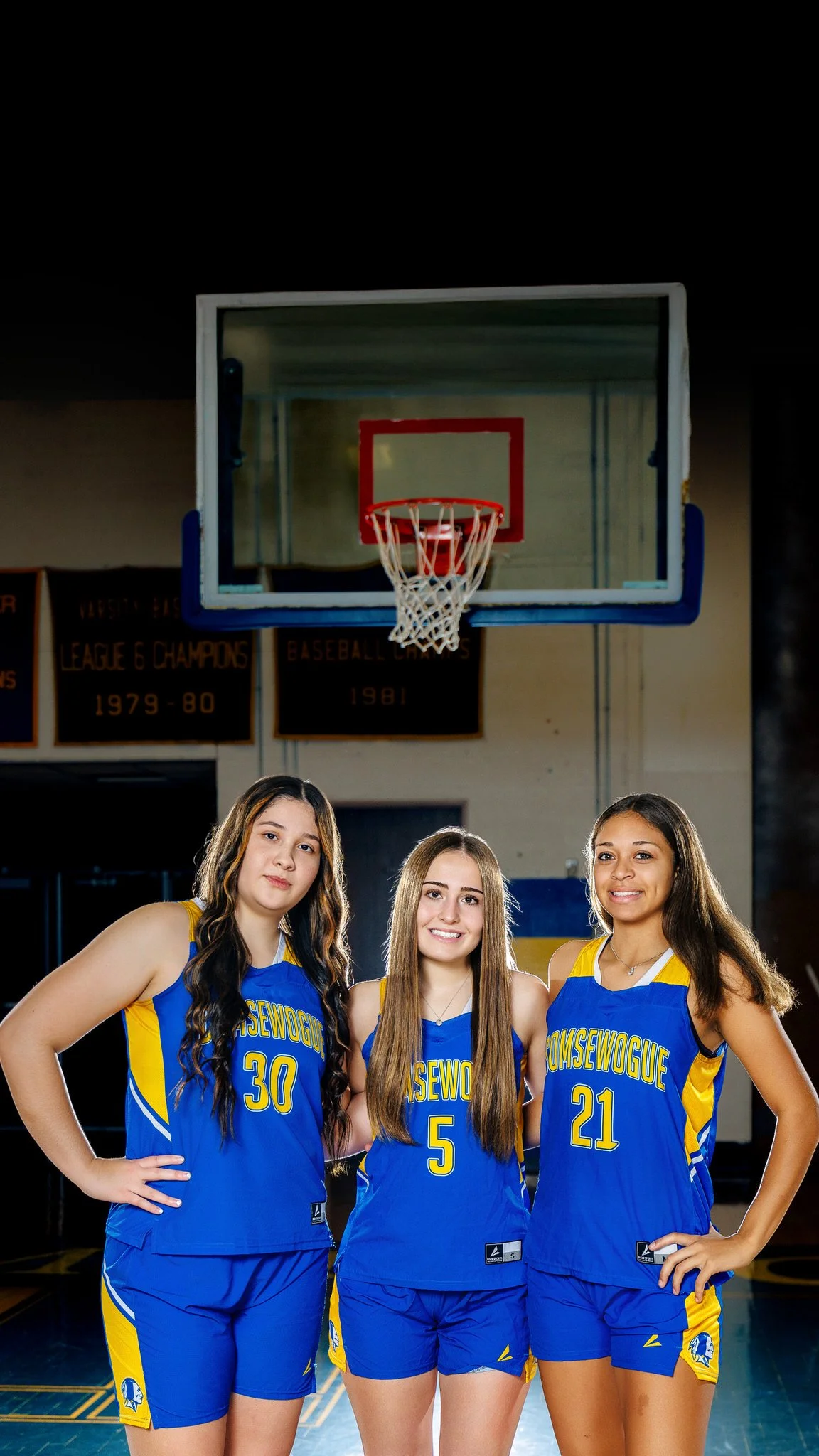 Three female basketball players in blue and yellow uniforms standing on a basketball court in front of a basketball hoop, smiling at the camera.