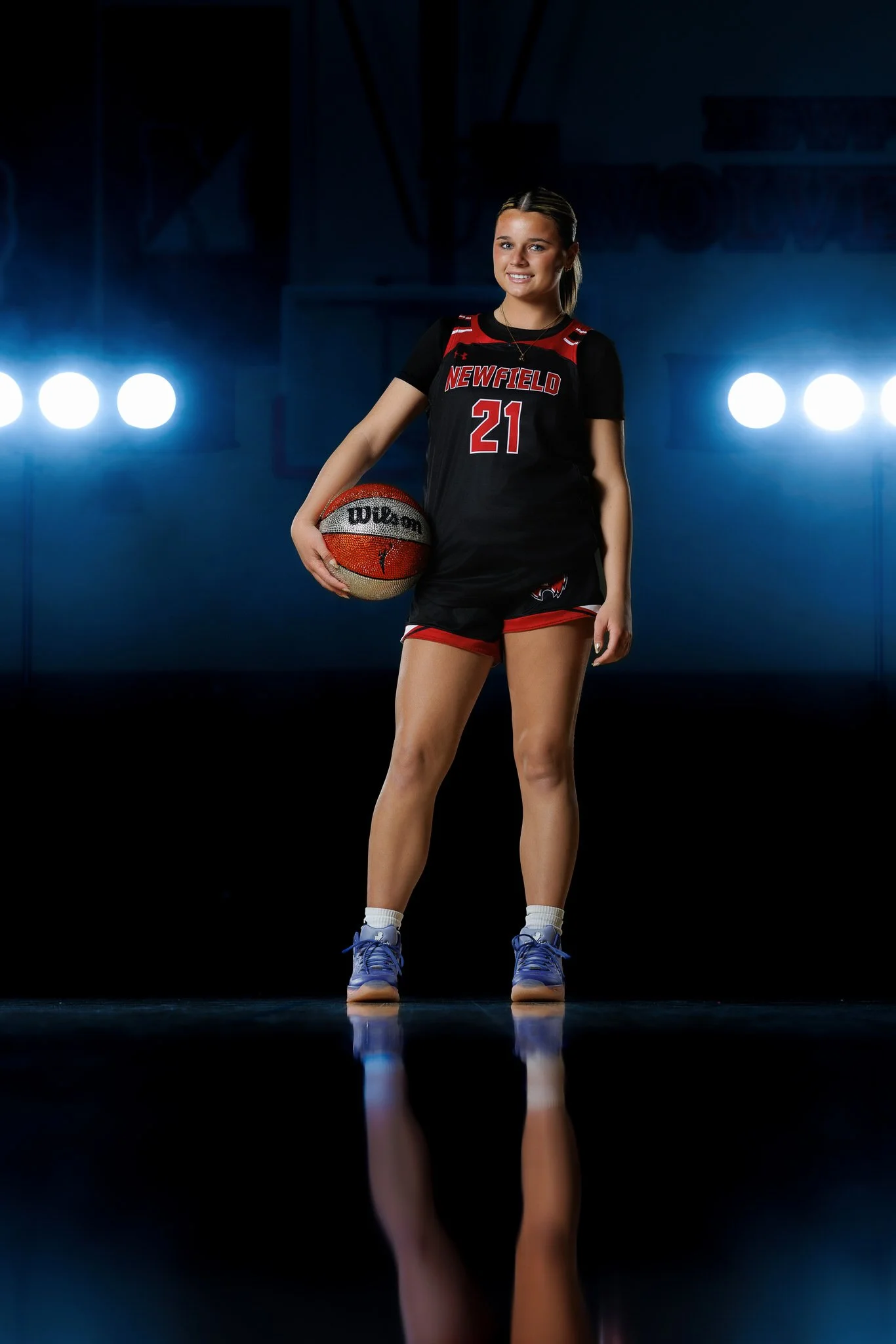 A young female basketball player in a black and red uniform, holding a basketball, standing on a court with bright lights behind her.
