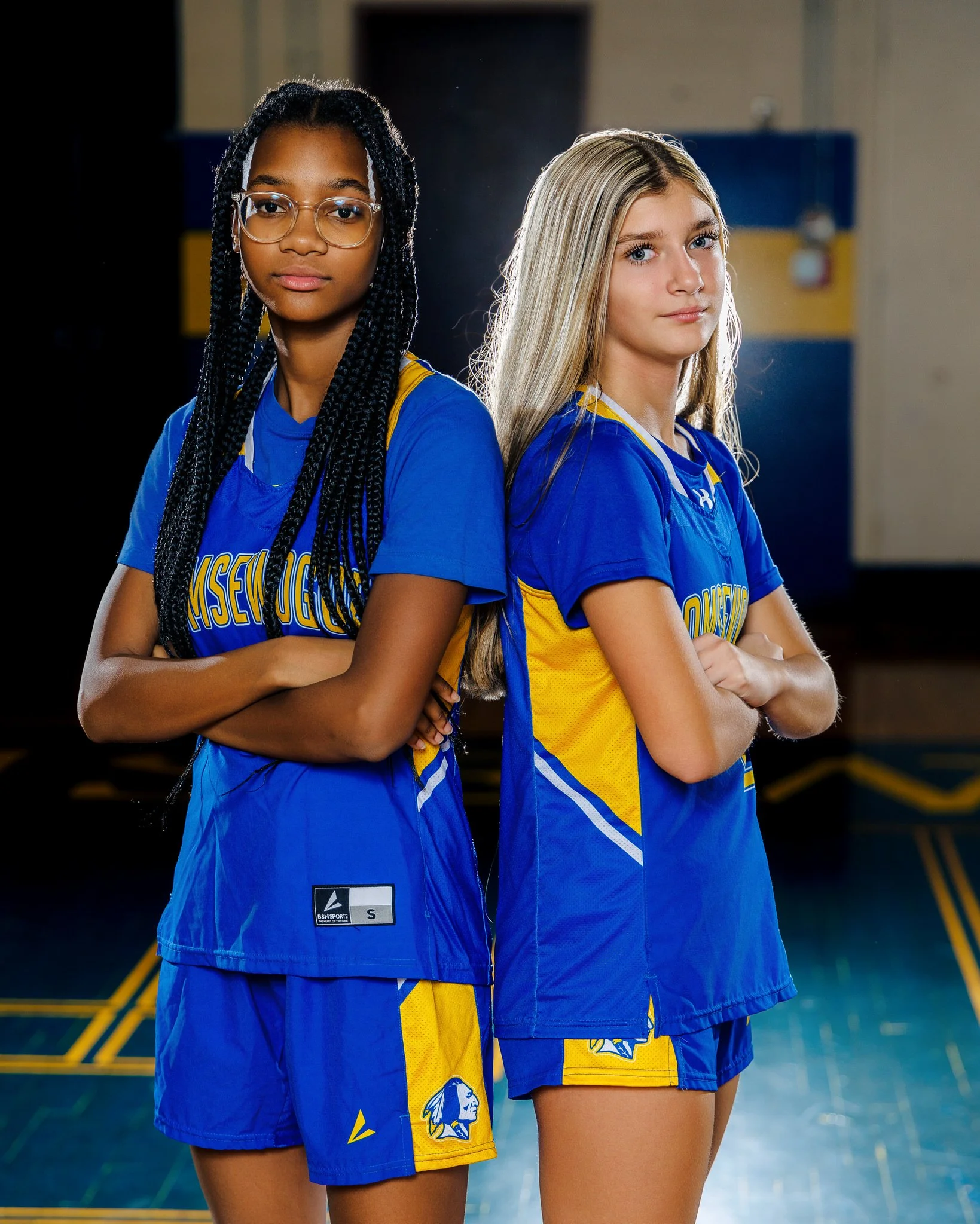 Two teenage girls in blue and yellow volleyball uniforms standing back-to-back with arms crossed in a gymnasium.