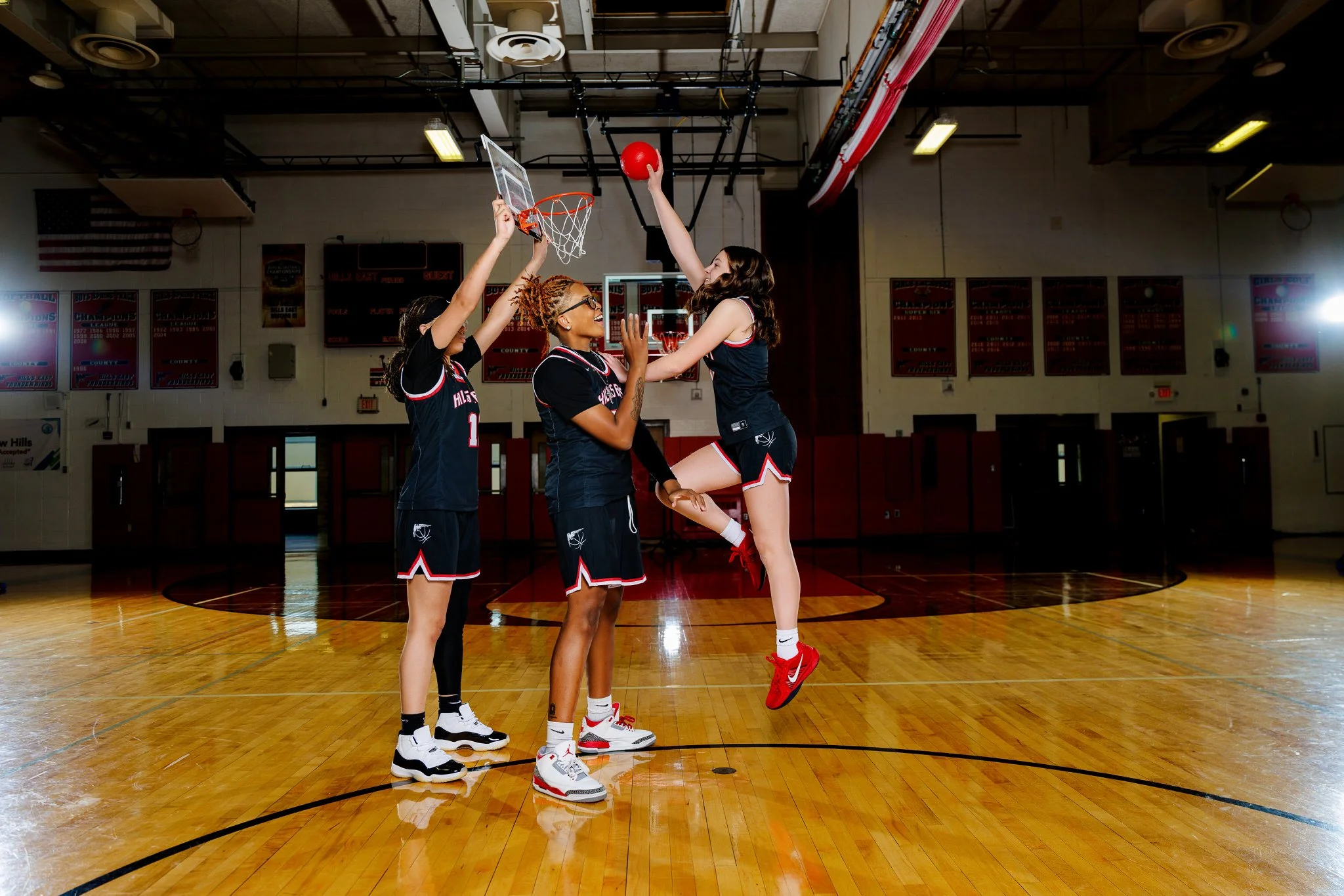 Four female basketball players practicing in an indoor gym. One player in a black jersey is jumping with the ball about to shoot at the hoop, while the three other players are defending.