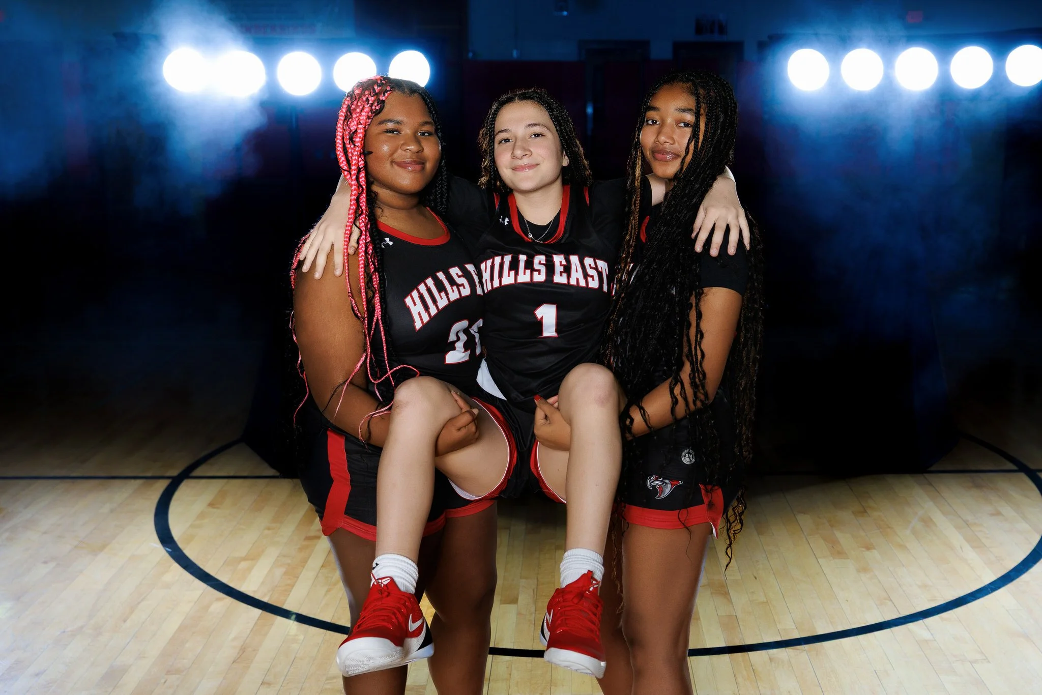 Three young female basketball players in black and red uniforms standing on a gymnasium court, with one girl being lifted by the other two. They are smiling and posing for the camera, with bright lights and a dark background behind them.