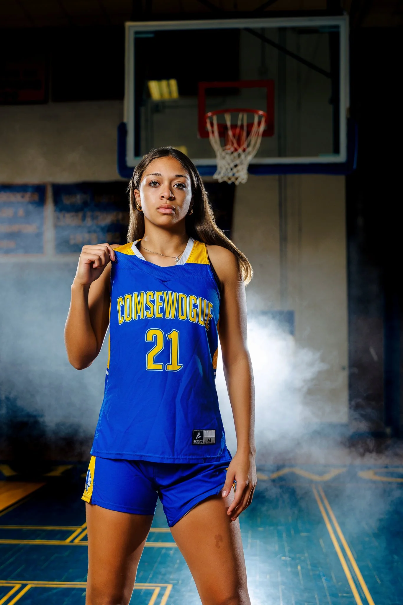 A young woman in a blue basketball uniform with yellow accents and the number 21, standing in a gymnasium in front of a basketball hoop, holding her jersey.