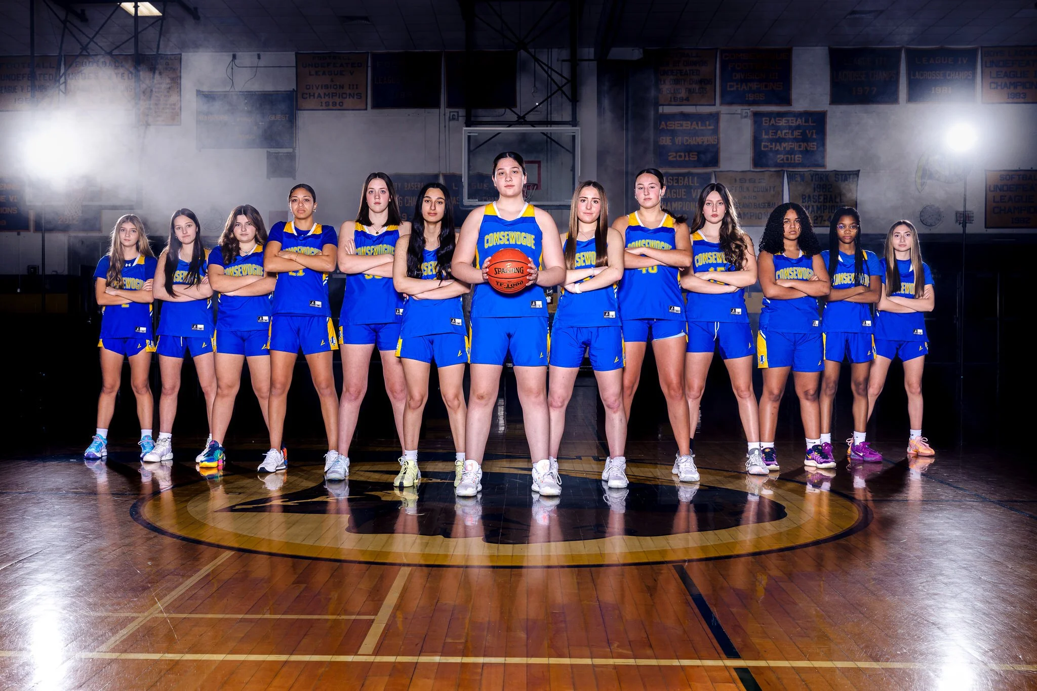 A girls' basketball team in blue uniforms standing in a gymnasium with a basketball at the center, formation in a line with arms crossed, under bright lights with banners hanging in the background.