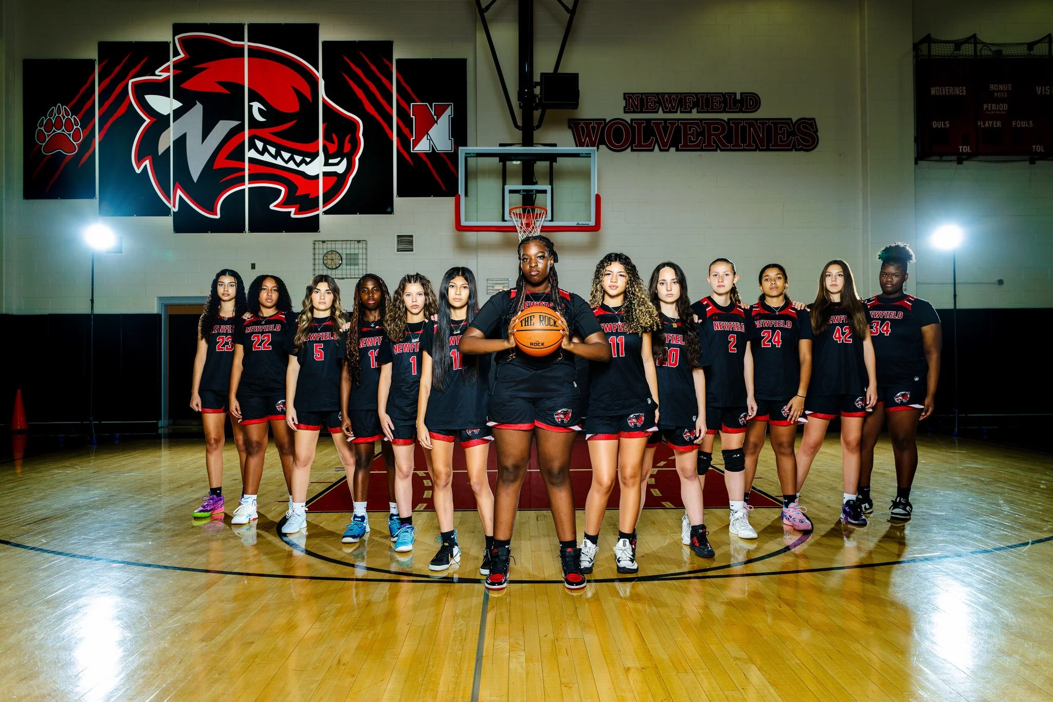 A girls basketball team in black and red uniforms standing on a basketball court with a coach holding a ball in the center, in a gymnasium.