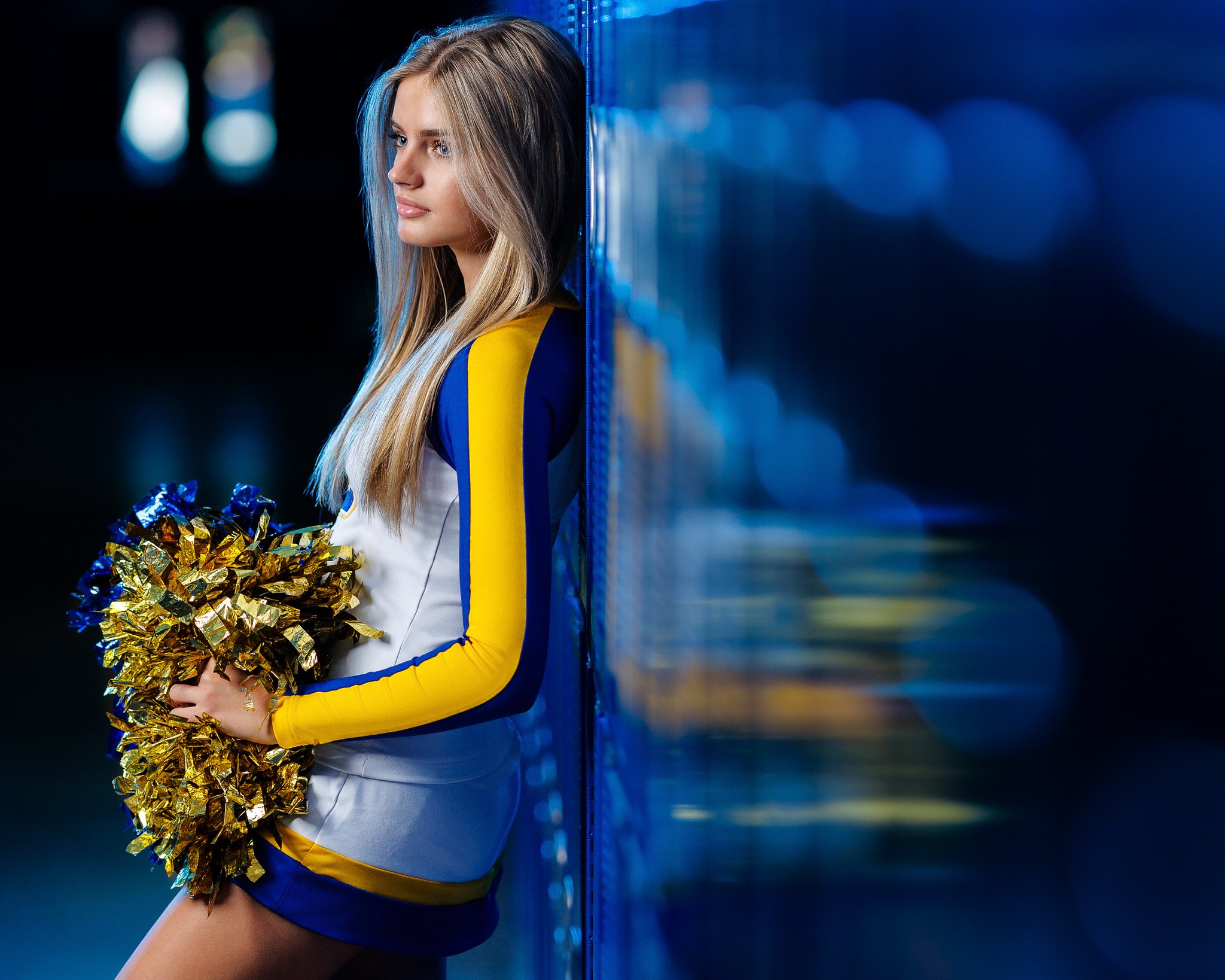 Cheerleader standing against a blue locker, holding gold and blue pom-poms, in a sports stadium at night.