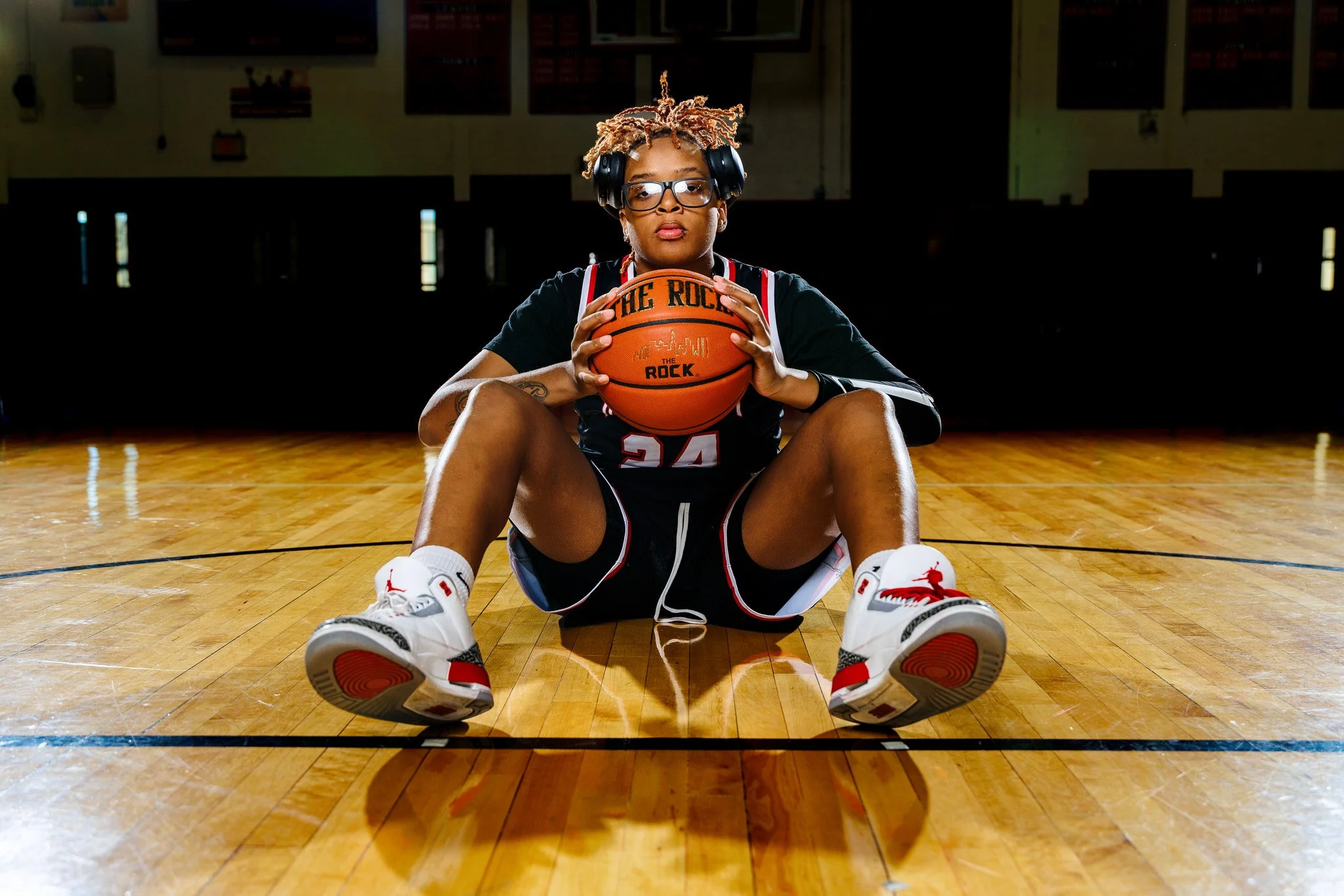 Female basketball player sitting on a gymnasium floor, holding a basketball, wearing a black jersey, shorts, and white sneakers with red accents, with headphones over her head and glasses.