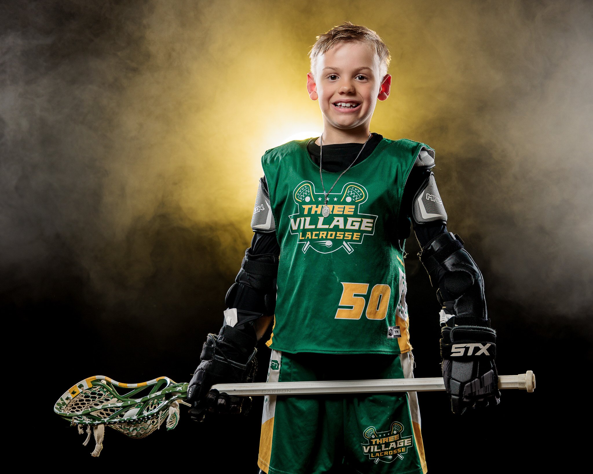 Young lacrosse player wearing "Three Village Lacrosse" uniform, holding a lacrosse stick, with protective gear, and standing against a smoky background.