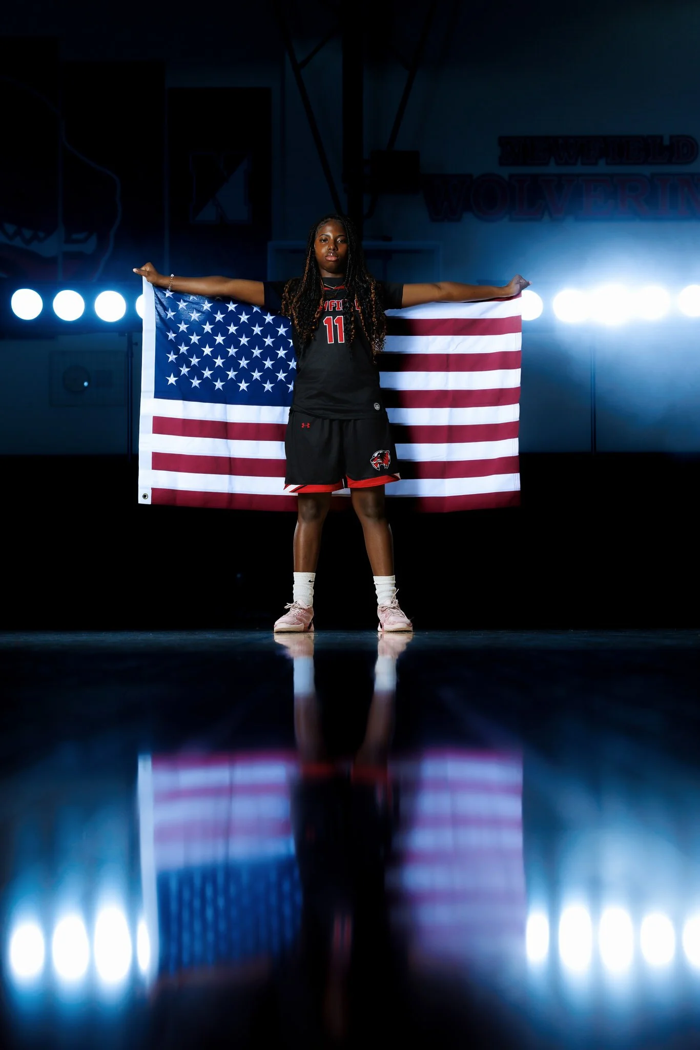 A female basketball player in a black uniform holding an American flag, standing under bright lights in an indoor basketball court.