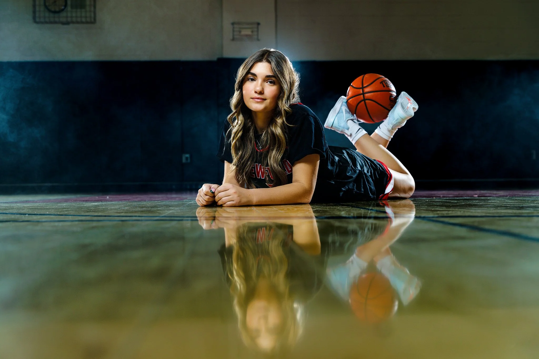 A young woman lying on her stomach on a basketball court with a basketball resting on her legs, in an indoor gym. She is looking at the camera with a relaxed expression, wearing a black T-shirt and athletic shorts.