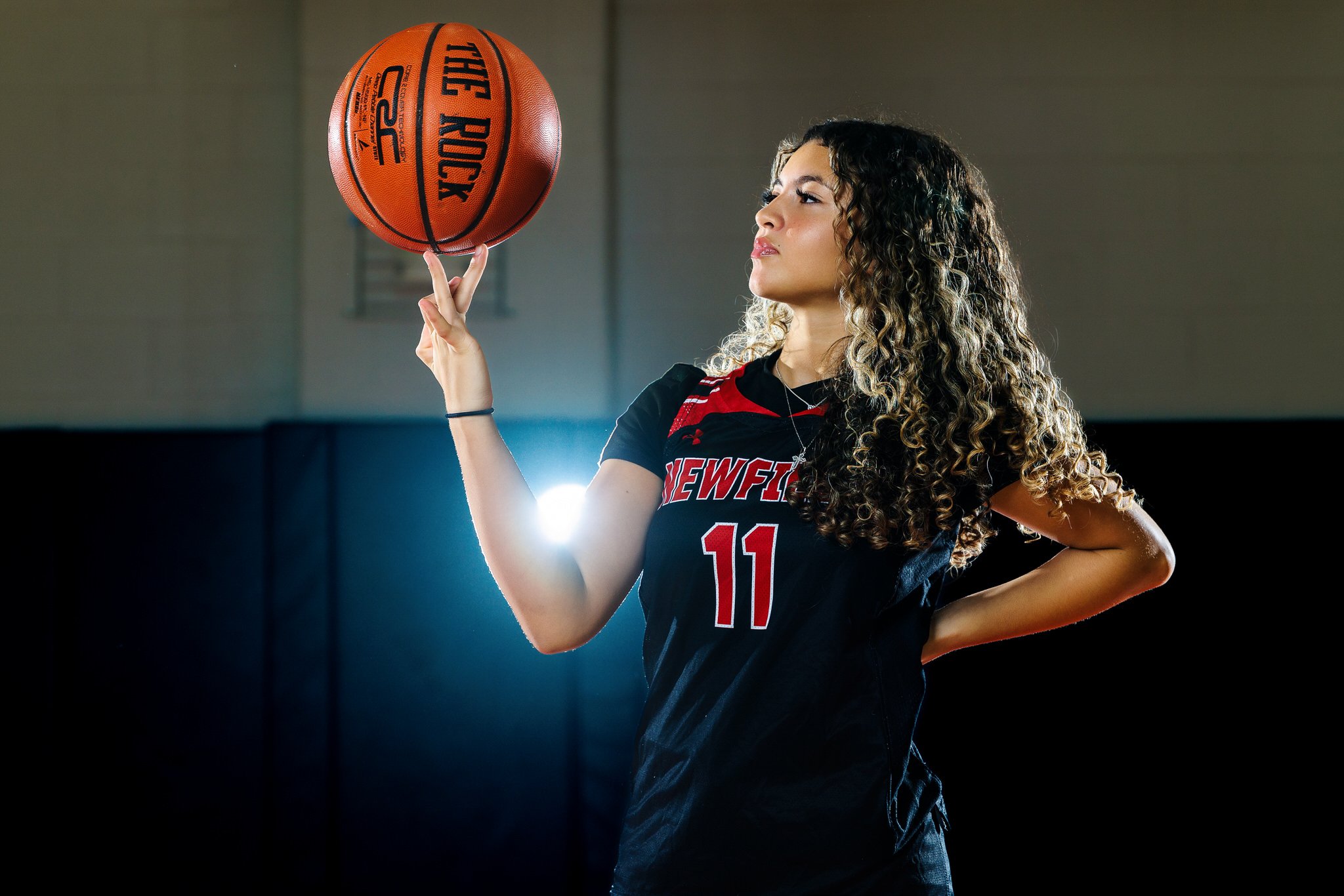 Young woman with curly hair in a black and red volleyball uniform spinning a basketball on her finger in a gymnasium.