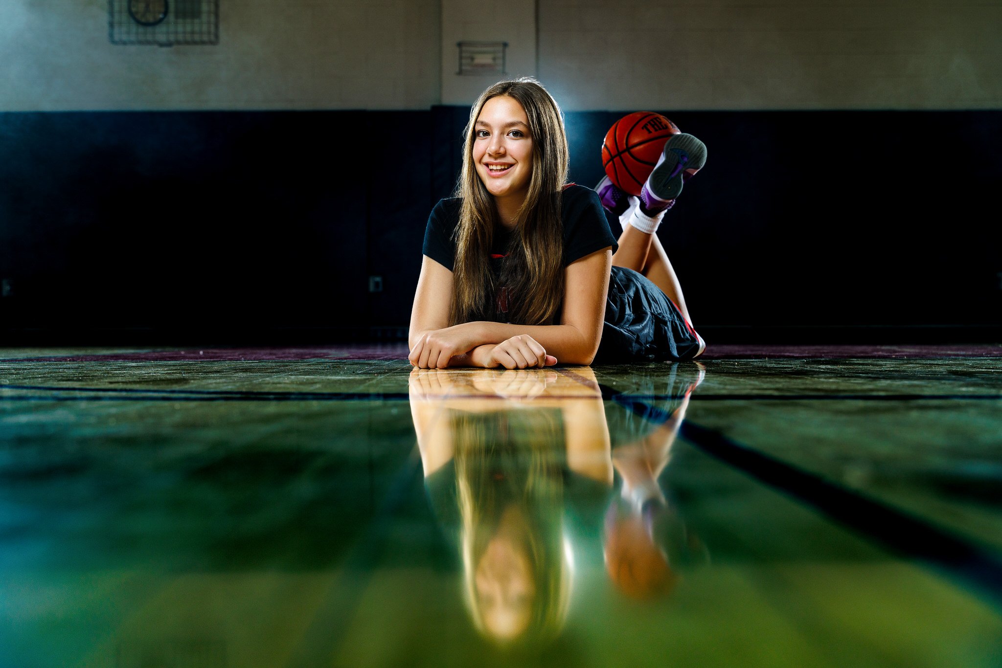 A young woman laying on her stomach on a gym floor, smiling, with her hands clasped in front of her. A basketball is behind her knees, and she is wearing athletic clothing and sneakers.