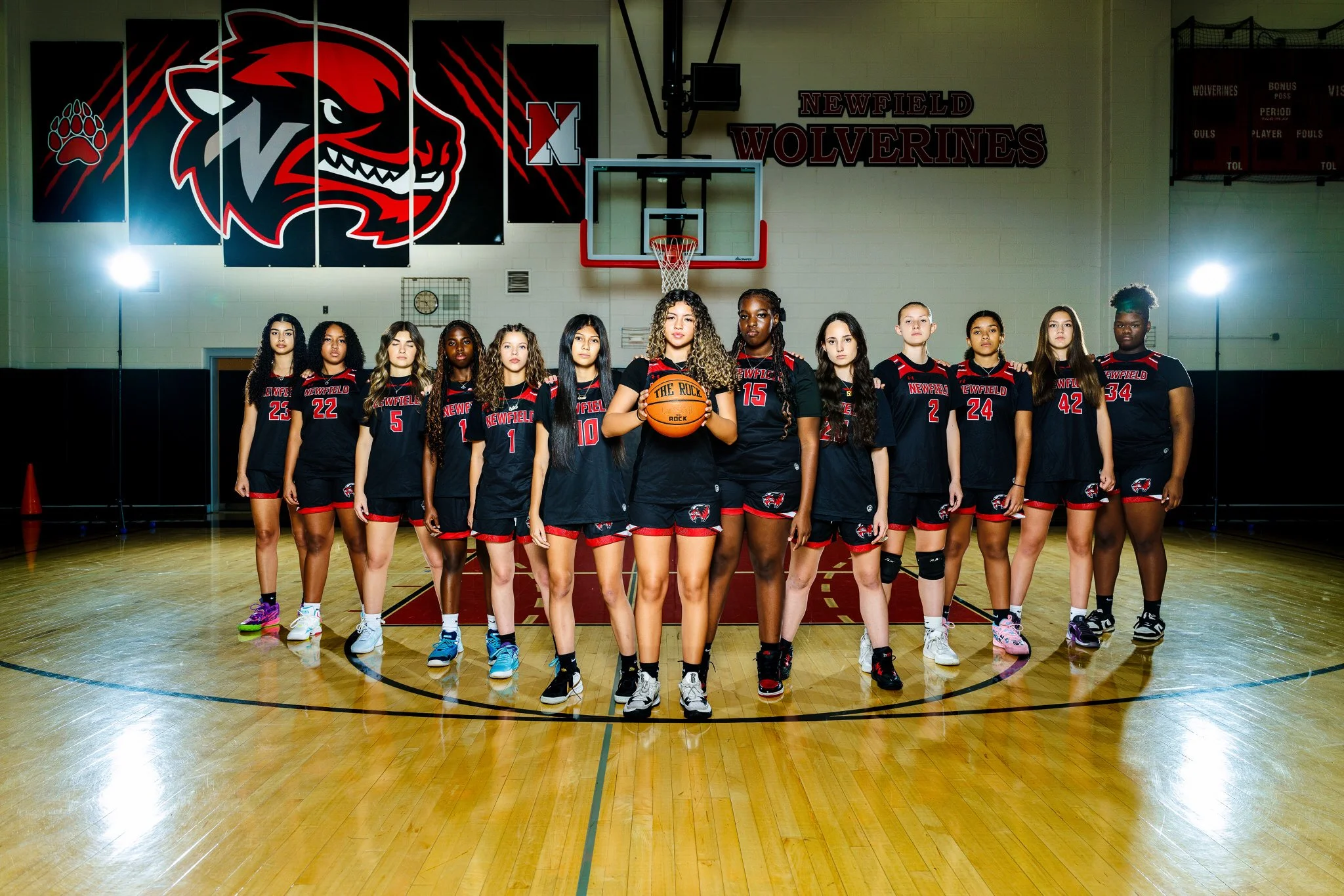 A girls' basketball team standing on a gymnasium basketball court in uniform, with the team holding a basketball and posing for a team photo in front of a basketball hoop and wall with 'Newfield Wolverines' signage.