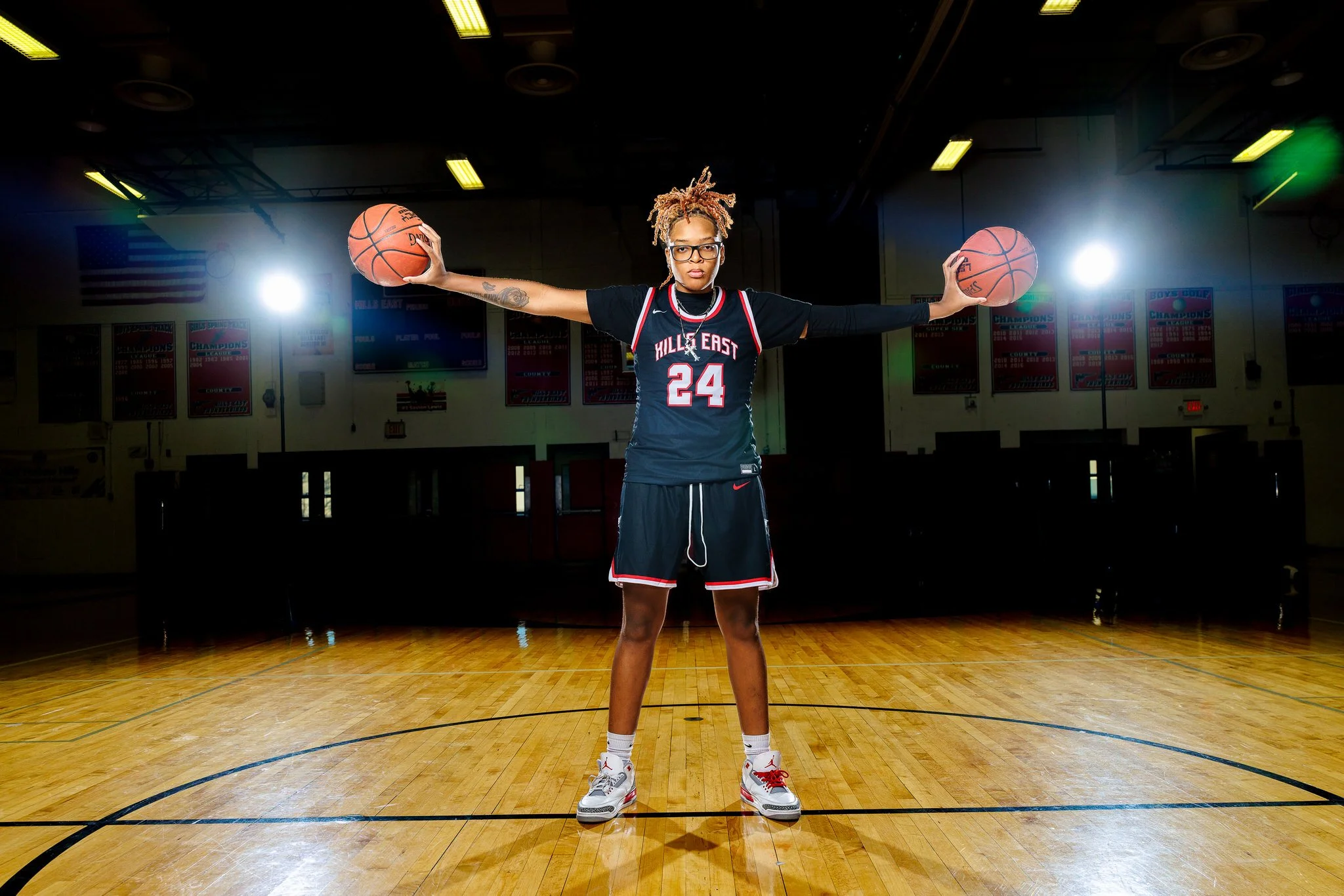A female basketball player stands on a basketball court with arms outstretched, holding basketballs in each hand, inside a gymnasium with banners hanging on the walls.
