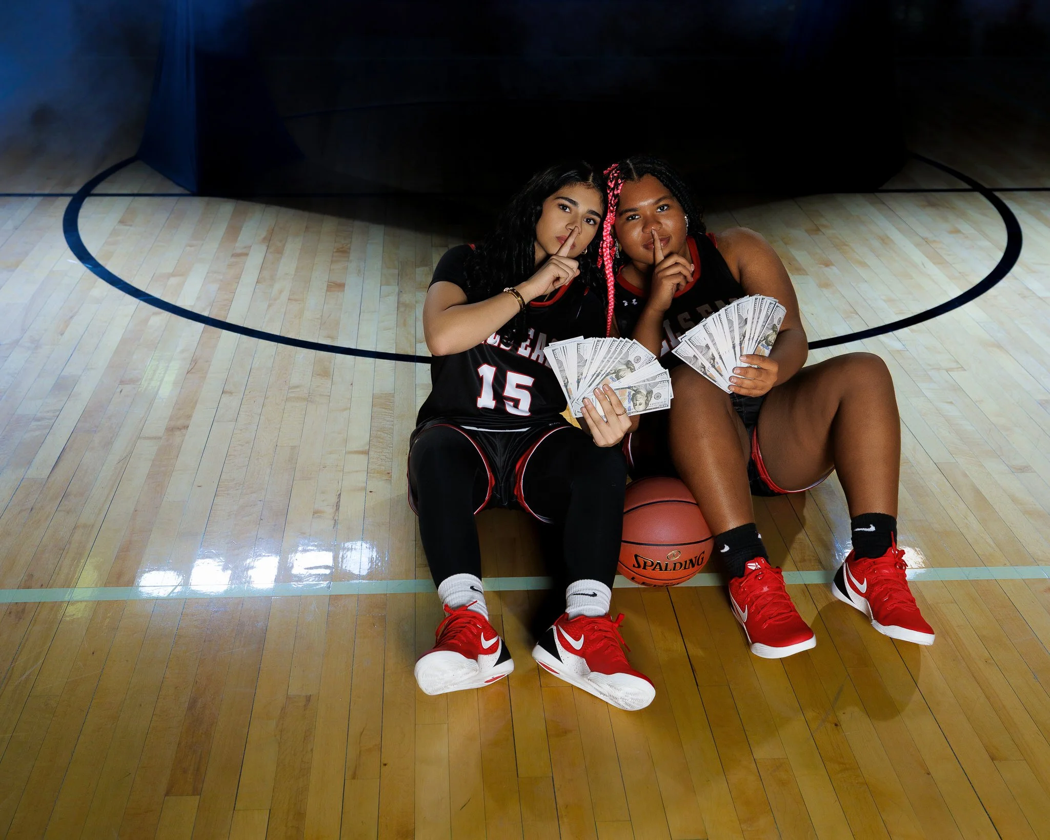 Two young female basketball players sitting on the gym floor with a basketball, holding fans of dollar bills, and making a shushing gesture with their fingers to their lips.