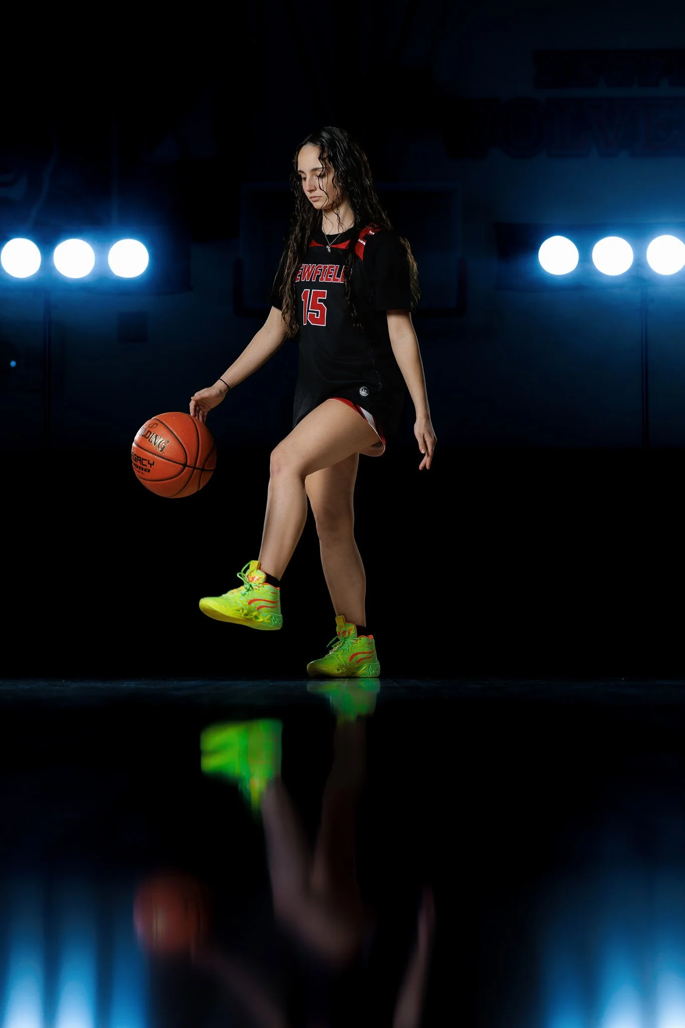 A female basketball player in a black uniform with red accents is performing a move while balancing a basketball on her left foot in a dark gym with bright lights behind her.
