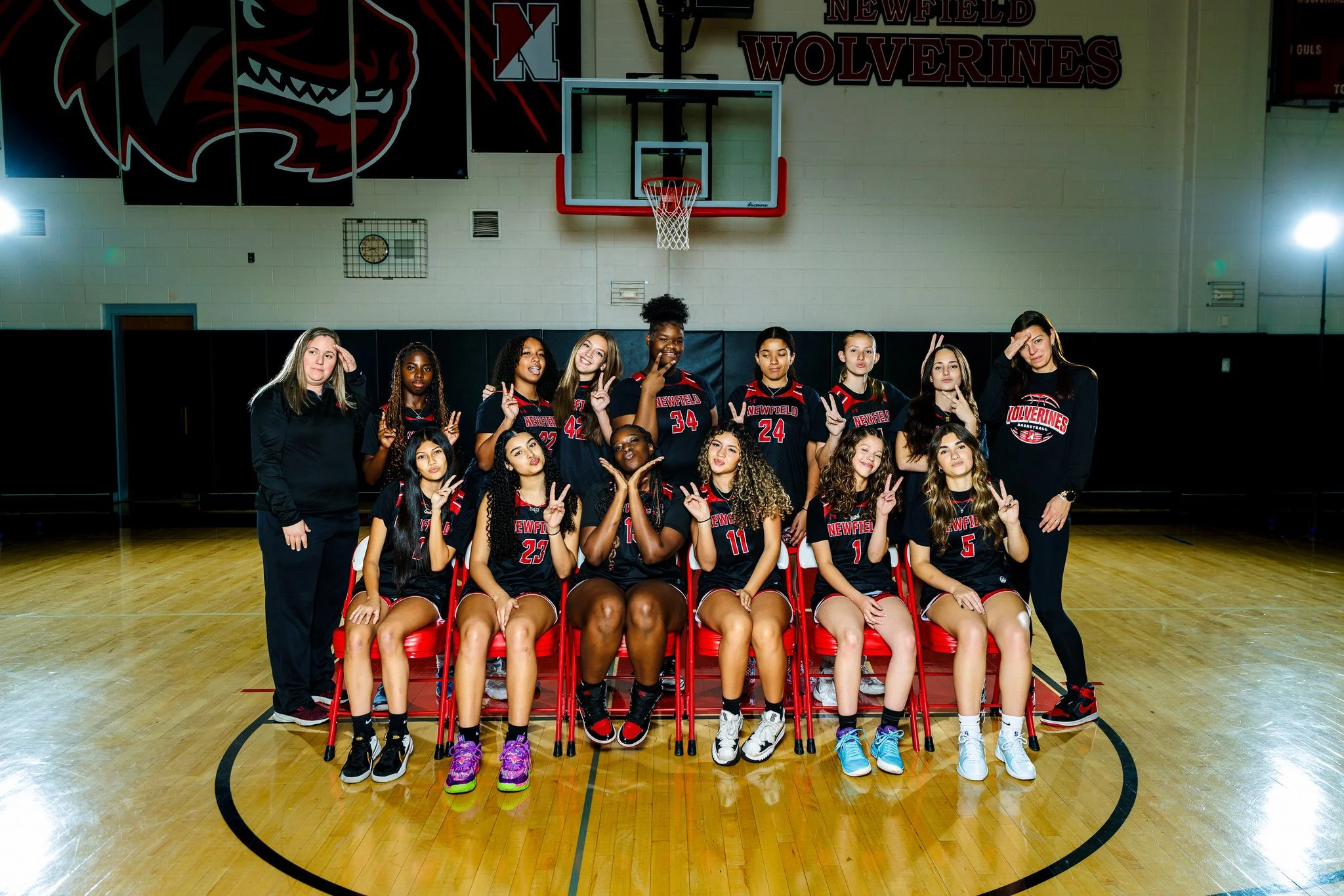 Group photo of a girls' basketball team in a gymnasium, with players in black and red uniforms and two coaches, some seated and some standing, with a basketball hoop and scoreboard in the background.