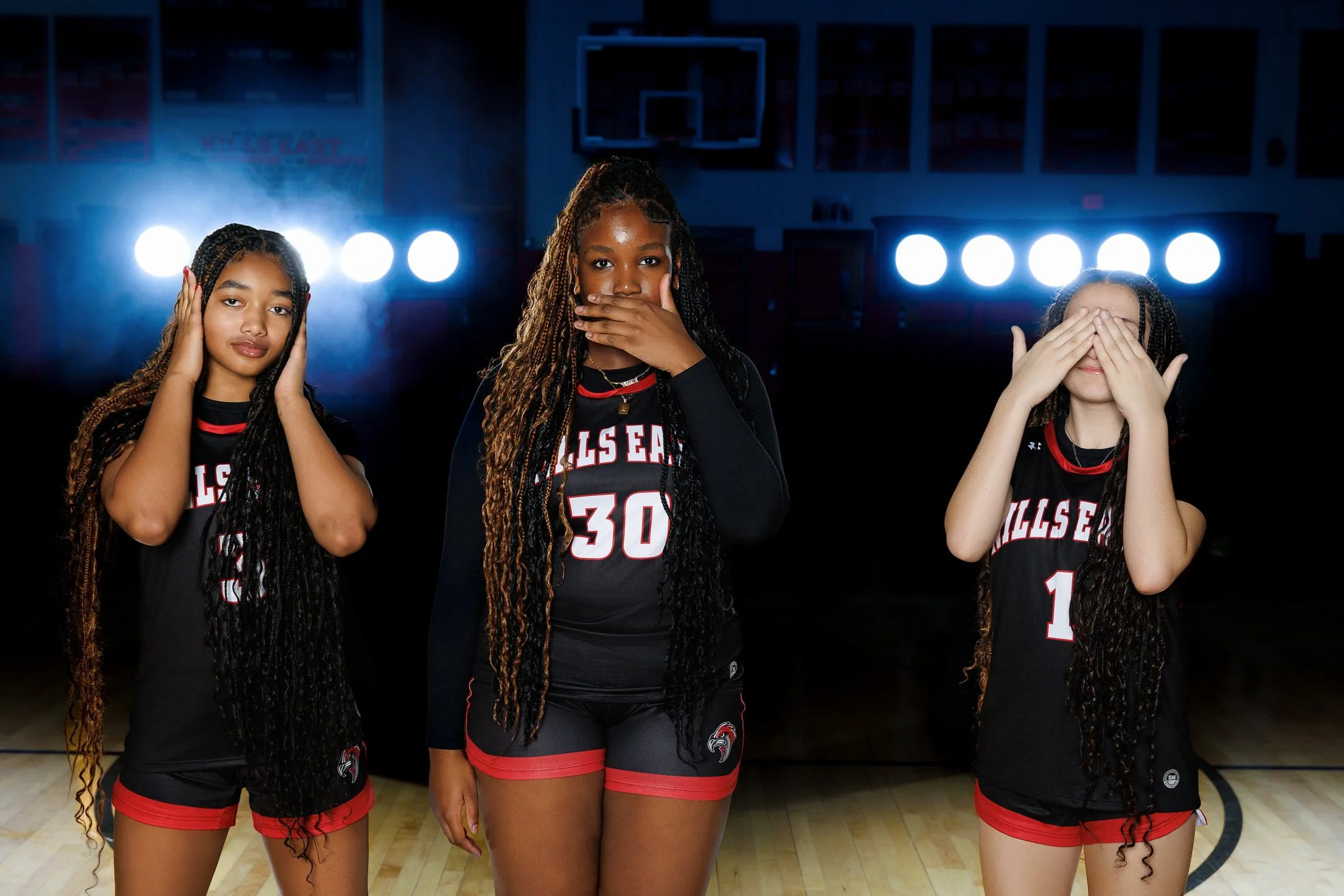 Three young female basketball players in black and red uniforms standing on a basketball court, with bright lights behind them.