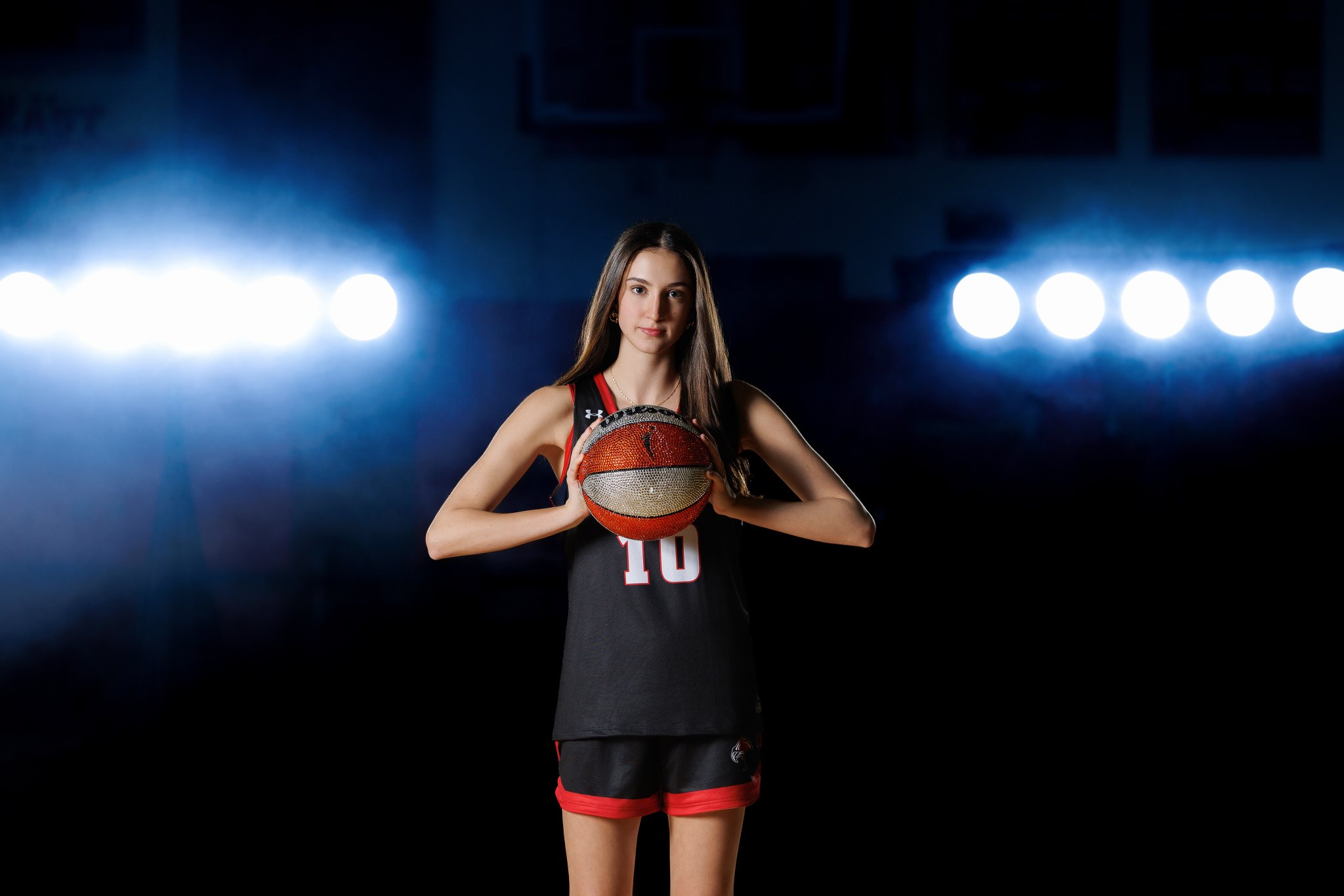 A young female basketball player in a black and red uniform holding a glittery basketball in a dark gym with bright lights in the background.